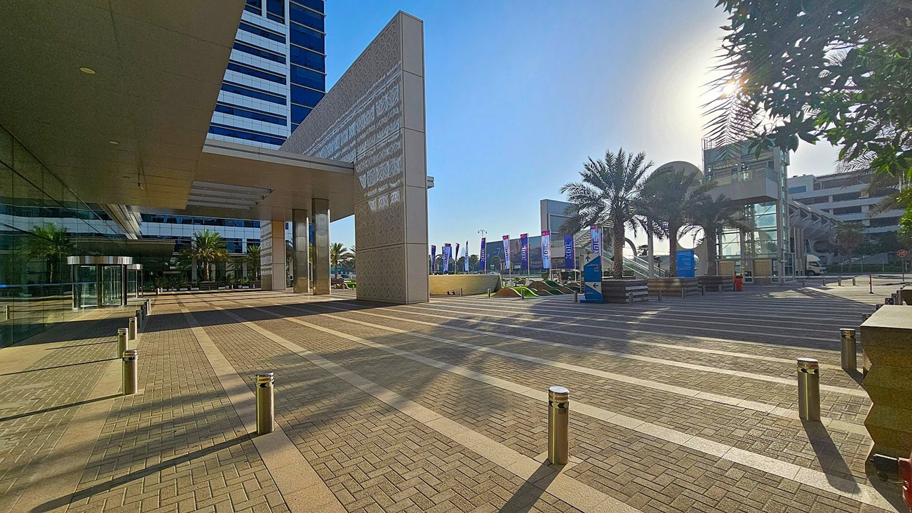 A modern urban plaza with a patterned pavement and metal bollards. On the left, a tall building with reflective glass windows and a covered walkway. In the centre, palm trees and a series of flags line the path. On the right, a structure with glass and metal elements, partially obscured by trees. The sun is visible through the trees, casting shadows on the ground. A modern urban plaza with a patterned pavement and metal bollards. On the left, a tall building with reflective glass windows and a covered walkway. In the centre, palm trees and a series of flags line the path. On the right, a structure with glass and metal elements, partially obscured by trees. The sun is visible through the trees, casting shadows on the ground.