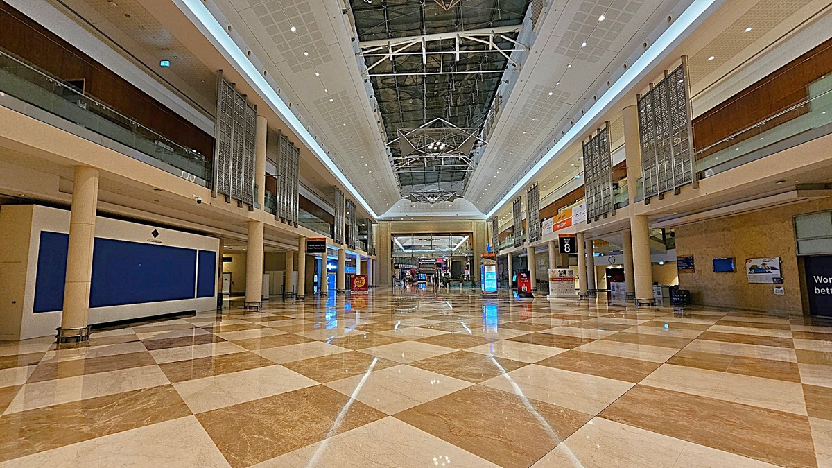 A spacious indoor area with a polished, chequered floor in shades of beige and brown. The ceiling is high with a geometric design and bright lighting. On both sides, there are two levels with glass railings and decorative panels. Various signs and digital displays are visible along the walls. In the background, there are more signs and a few people walking. A spacious indoor area with a polished, chequered floor in shades of beige and brown. The ceiling is high with a geometric design and bright lighting. On both sides, there are two levels with glass railings and decorative panels. Various signs and digital displays are visible along the walls. In the background, there are more signs and a few people walking.