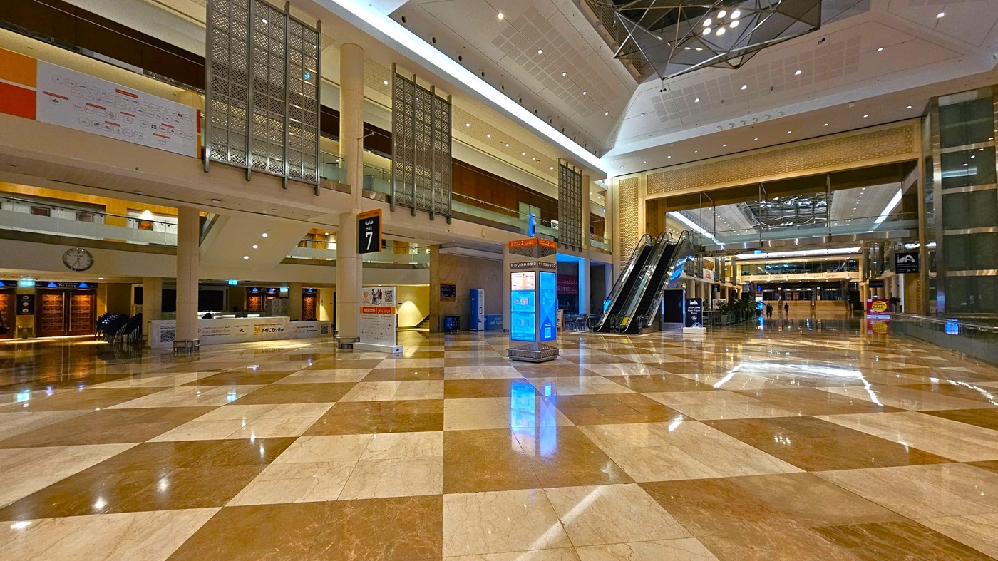 A spacious indoor area with a polished, chequered marble floor. On the left, there are information desks and a large wall clock. In the centre, a digital information kiosk stands near escalators leading to an upper level. The ceiling is high with modern lighting fixtures. The background features a large open space with more seating and signage. The area is well-lit and appears to be part of a public or commercial building. A spacious indoor area with a polished, chequered marble floor. On the left, there are information desks and a large wall clock. In the centre, a digital information kiosk stands near escalators leading to an upper level. The ceiling is high with modern lighting fixtures. The background features a large open space with more seating and signage. The area is well-lit and appears to be part of a public or commercial building.