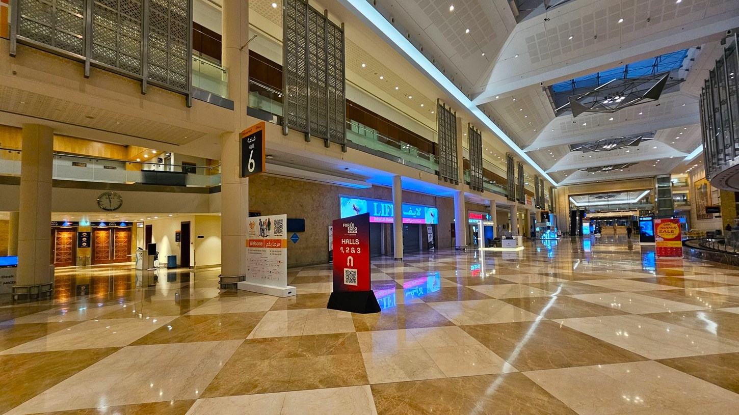 A spacious indoor hall with a polished, reflective marble floor in a checkered pattern of beige and brown. On the left, a large "WELCOME" sign in English and Arabic is displayed above a reception area. A digital kiosk with bright screens stands in the centre. On the right, a wide staircase leads to an upper level with glass railings. The ceiling is high with geometric designs and recessed lighting. Hall 8 is indicated by a sign hanging from the ceiling. A spacious indoor hall with a polished, reflective marble floor in a checkered pattern of beige and brown. On the left, a large "WELCOME" sign in English and Arabic is displayed above a reception area. A digital kiosk with bright screens stands in the centre. On the right, a wide staircase leads to an upper level with glass railings. The ceiling is high with geometric designs and recessed lighting. Hall 8 is indicated by a sign hanging from the ceiling.
