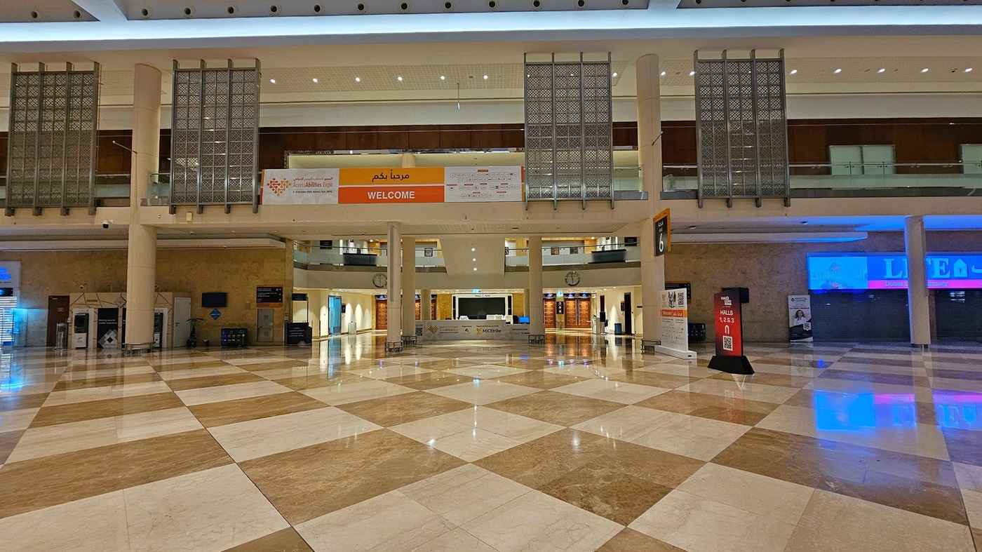 A spacious indoor hall with a polished, reflective floor featuring a diamond pattern in shades of beige and brown. On the left, there are signs for Hall 6 and Forex Expo, along with a clock and elevators. The right side has a "Super Sale" sign and various illuminated displays. The ceiling is high with geometric designs and bright lighting. The area is lined with shops and advertisements, and there are few people in the distance. A spacious indoor hall with a polished, reflective floor featuring a diamond pattern in shades of beige and brown. On the left, there are signs for Hall 6 and Forex Expo, along with a clock and elevators. The right side has a "Super Sale" sign and various illuminated displays. The ceiling is high with geometric designs and bright lighting. The area is lined with shops and advertisements, and there are few people in the distance.
