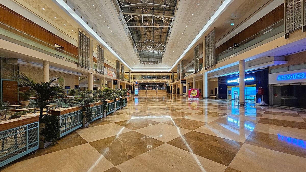 A spacious indoor area with a polished, chequered floor in shades of beige and brown. The ceiling is high with recessed lighting. In the centre, a large welcome banner is displayed above a reception desk. On the left, vending machines and a small seating area are visible. On the right, digital screens and signage for halls are present. The upper level features decorative panels and glass railings. A spacious indoor area with a polished, chequered floor in shades of beige and brown. The ceiling is high with recessed lighting. In the centre, a large welcome banner is displayed above a reception desk. On the left, vending machines and a small seating area are visible. On the right, digital screens and signage for halls are present. The upper level features decorative panels and glass railings.