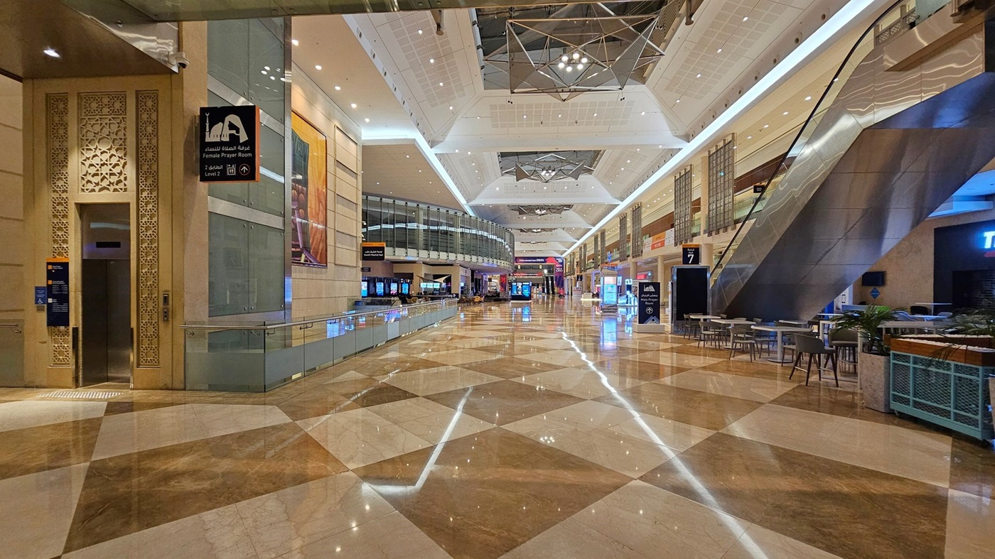 A spacious indoor area with a polished, reflective floor featuring a geometric pattern. On the left, there's a sign for a female prayer room next to an intricately designed doorway. In the centre, a wide corridor extends with various shops and digital displays. On the right, an escalator leads to an upper level, with tables and chairs nearby. The ceiling is high with modern lighting fixtures and geometric designs. A spacious indoor area with a polished, reflective floor featuring a geometric pattern. On the left, there's a sign for a female prayer room next to an intricately designed doorway. In the centre, a wide corridor extends with various shops and digital displays. On the right, an escalator leads to an upper level, with tables and chairs nearby. The ceiling is high with modern lighting fixtures and geometric designs.