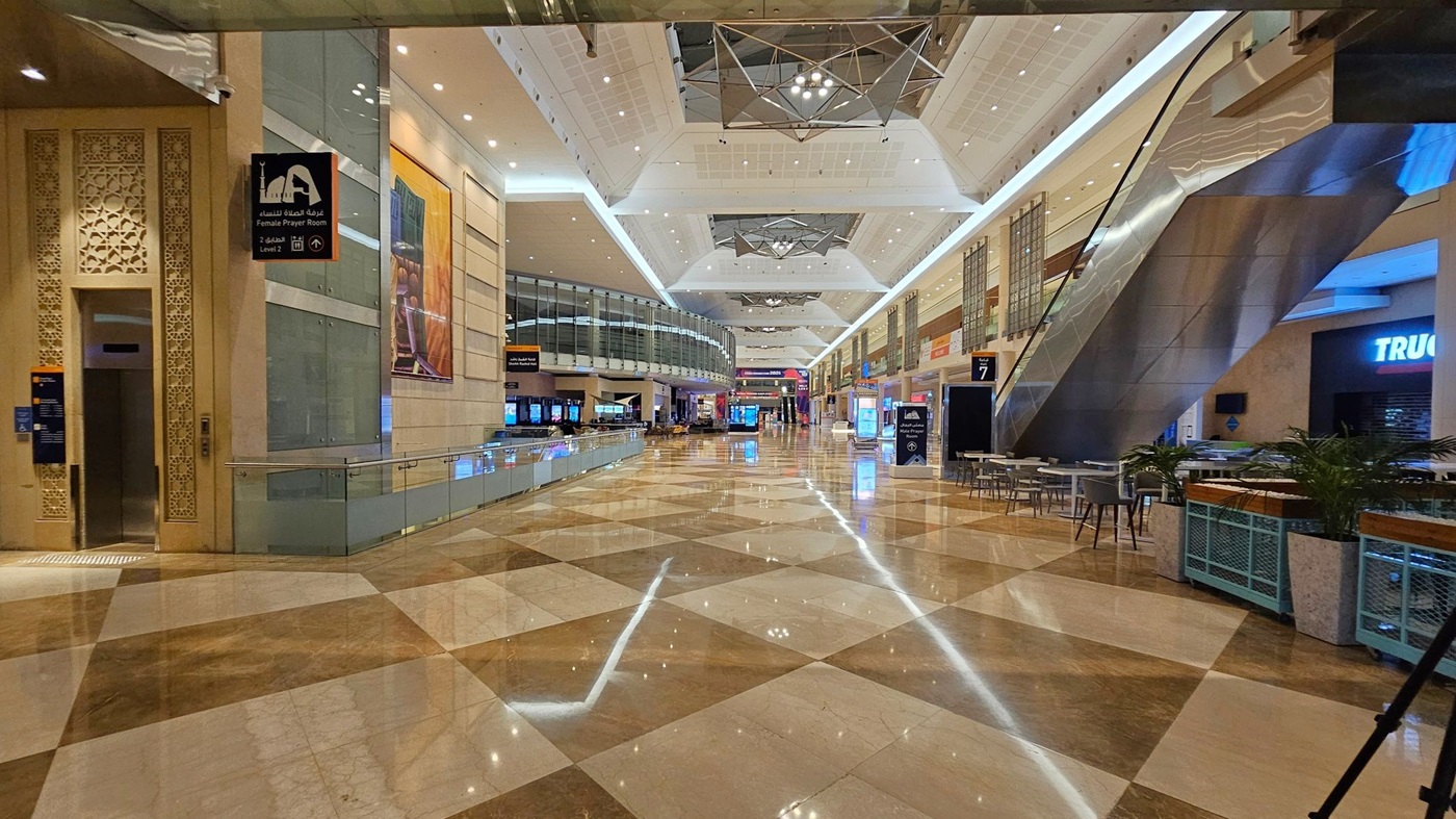 A spacious indoor area with a polished, reflective floor featuring a geometric pattern. On the left, there's a sign for a female prayer room and an intricately designed wall. The centre has a glass railing and a curved glass structure. On the right, there are escalators, seating areas with tables and chairs, and a shop with a partially visible sign. The ceiling is high with geometric light fixtures and recessed lighting. A spacious indoor area with a polished, reflective floor featuring a geometric pattern. On the left, there's a sign for a female prayer room and an intricately designed wall. The centre has a glass railing and a curved glass structure. On the right, there are escalators, seating areas with tables and chairs, and a shop with a partially visible sign. The ceiling is high with geometric light fixtures and recessed lighting.
