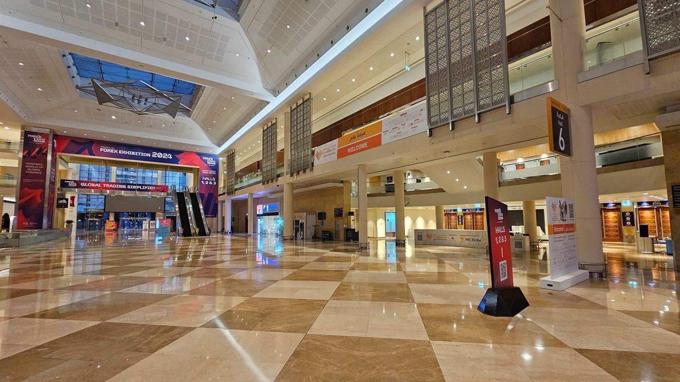 A spacious exhibition hall with a polished, reflective marble floor in a checkered pattern. On the left, a large banner reads "Forex Exhibition 2024" above an entrance with glass doors. Two escalators are visible nearby. On the right, a sign indicates Hall 6, and there are various informational signs and banners. The ceiling is high with a geometric design and a skylight. The upper level features decorative panels and a "Welcome" banner. A spacious exhibition hall with a polished, reflective marble floor in a checkered pattern. On the left, a large banner reads "Forex Exhibition 2024" above an entrance with glass doors. Two escalators are visible nearby. On the right, a sign indicates Hall 6, and there are various informational signs and banners. The ceiling is high with a geometric design and a skylight. The upper level features decorative panels and a "Welcome" banner.