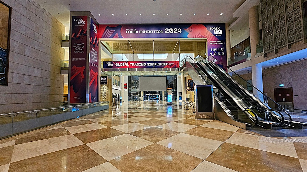 A spacious indoor area with a polished, chequered marble floor. On the left, there is a large wall with Forex Expo banners. In the centre, a wide entrance with a banner reading "Global Trading Simplified" leads to a hall. On the right, an escalator ascends to an upper level. The ceiling is high with recessed lighting, and the walls are adorned with additional banners and decorative panels. A spacious indoor area with a polished, chequered marble floor. On the left, there is a large wall with Forex Expo banners. In the centre, a wide entrance with a banner reading "Global Trading Simplified" leads to a hall. On the right, an escalator ascends to an upper level. The ceiling is high with recessed lighting, and the walls are adorned with additional banners and decorative panels.