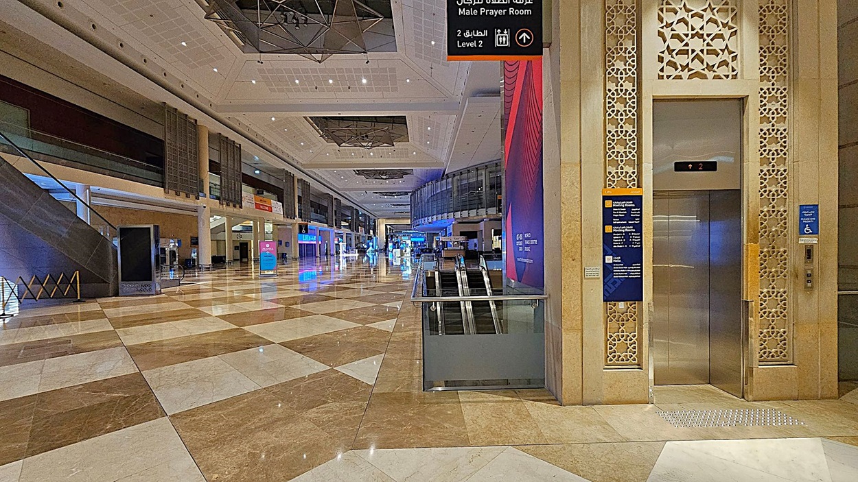 A spacious indoor area with a polished, patterned floor of brown and white tiles. On the right, there's an elevator with intricate decorative panels and signage indicating directions. In the centre, an escalator descends. The left side features a staircase and various signs. The ceiling is high with geometric designs and lighting fixtures. The background shows a long corridor with illuminated signs and displays. A spacious indoor area with a polished, patterned floor of brown and white tiles. On the right, there's an elevator with intricate decorative panels and signage indicating directions. In the centre, an escalator descends. The left side features a staircase and various signs. The ceiling is high with geometric designs and lighting fixtures. The background shows a long corridor with illuminated signs and displays.