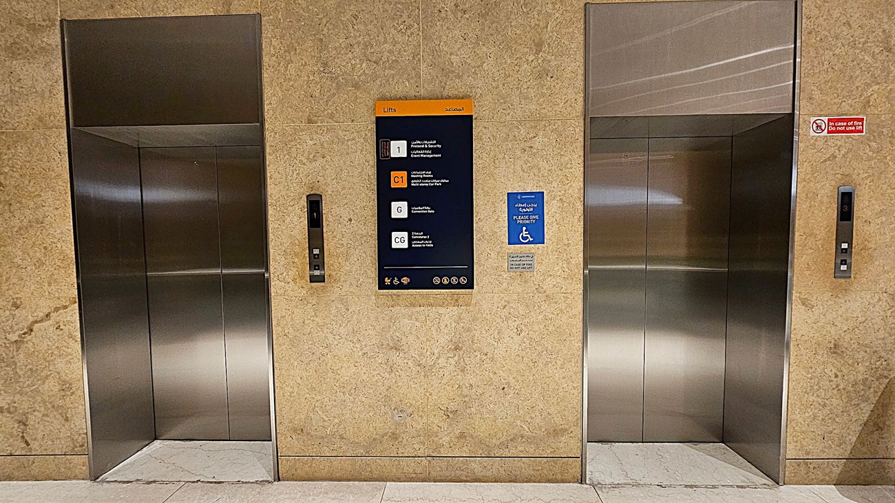 Two stainless steel lift doors are set into a beige marble wall. In the centre, a sign displays floor information in multiple languages. To the right of the sign, a blue notice indicates priority for wheelchair users. Each lift has a control panel with buttons and floor indicators. The floor is light-coloured stone, and the ceiling is not visible. Two stainless steel lift doors are set into a beige marble wall. In the centre, a sign displays floor information in multiple languages. To the right of the sign, a blue notice indicates priority for wheelchair users. Each lift has a control panel with buttons and floor indicators. The floor is light-coloured stone, and the ceiling is not visible.