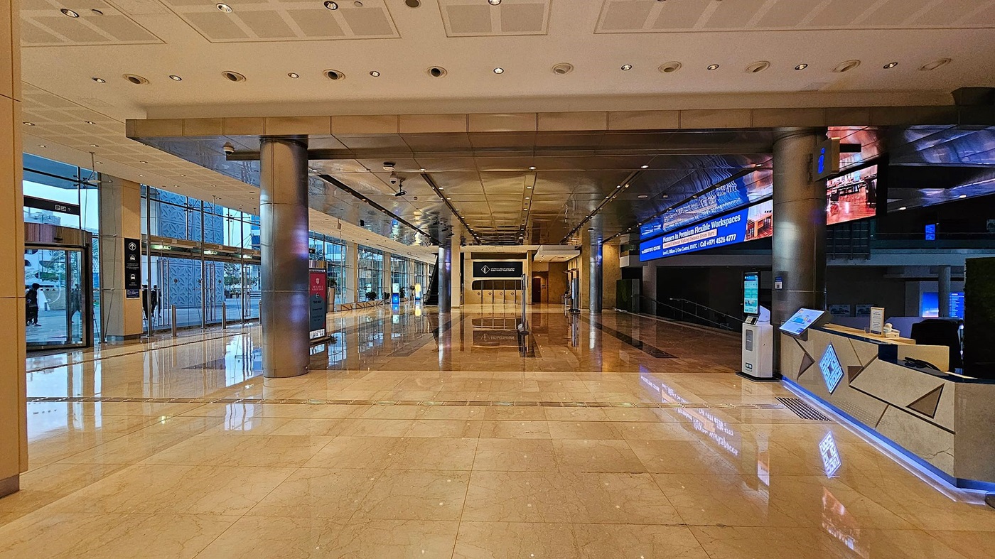 A spacious, modern lobby with a shiny marble floor and a grid-patterned ceiling with recessed lighting. On the left, there are dark reflective surfaces and a reception area. The right side features large glass windows with views of an outdoor area, including palm trees and banners. In the centre, there are glass doors and a large pillar. The space is well-lit, with reflections visible on the floor A spacious, modern lobby with a shiny marble floor and a grid-patterned ceiling with recessed lighting. On the left, there are dark reflective surfaces and a reception area. The right side features large glass windows with views of an outdoor area, including palm trees and banners. In the centre, there are glass doors and a large pillar. The space is well-lit, with reflections visible on the floor