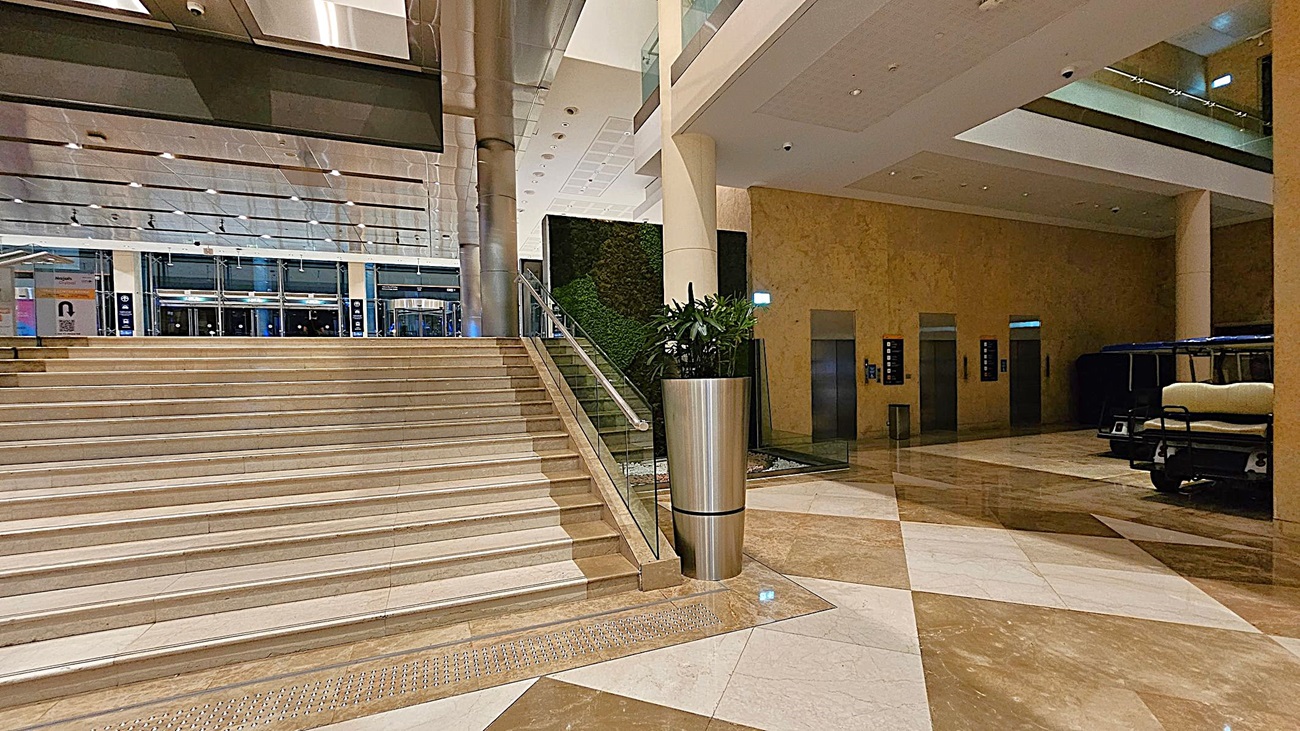 A spacious indoor area with a wide staircase on the left, featuring glass railings. In the centre, a large metal planter with green plants stands next to the stairs. The background shows a wall with a green vertical garden. On the right, there are several lifts with illuminated indicators and a small vehicle parked nearby. The floor is polished with a geometric pattern of light and dark tiles, and the ceiling is fitted with recessed lighting. A spacious indoor area with a wide staircase on the left, featuring glass railings. In the centre, a large metal planter with green plants stands next to the stairs. The background shows a wall with a green vertical garden. On the right, there are several lifts with illuminated indicators and a small vehicle parked nearby. The floor is polished with a geometric pattern of light and dark tiles, and the ceiling is fitted with recessed lighting.