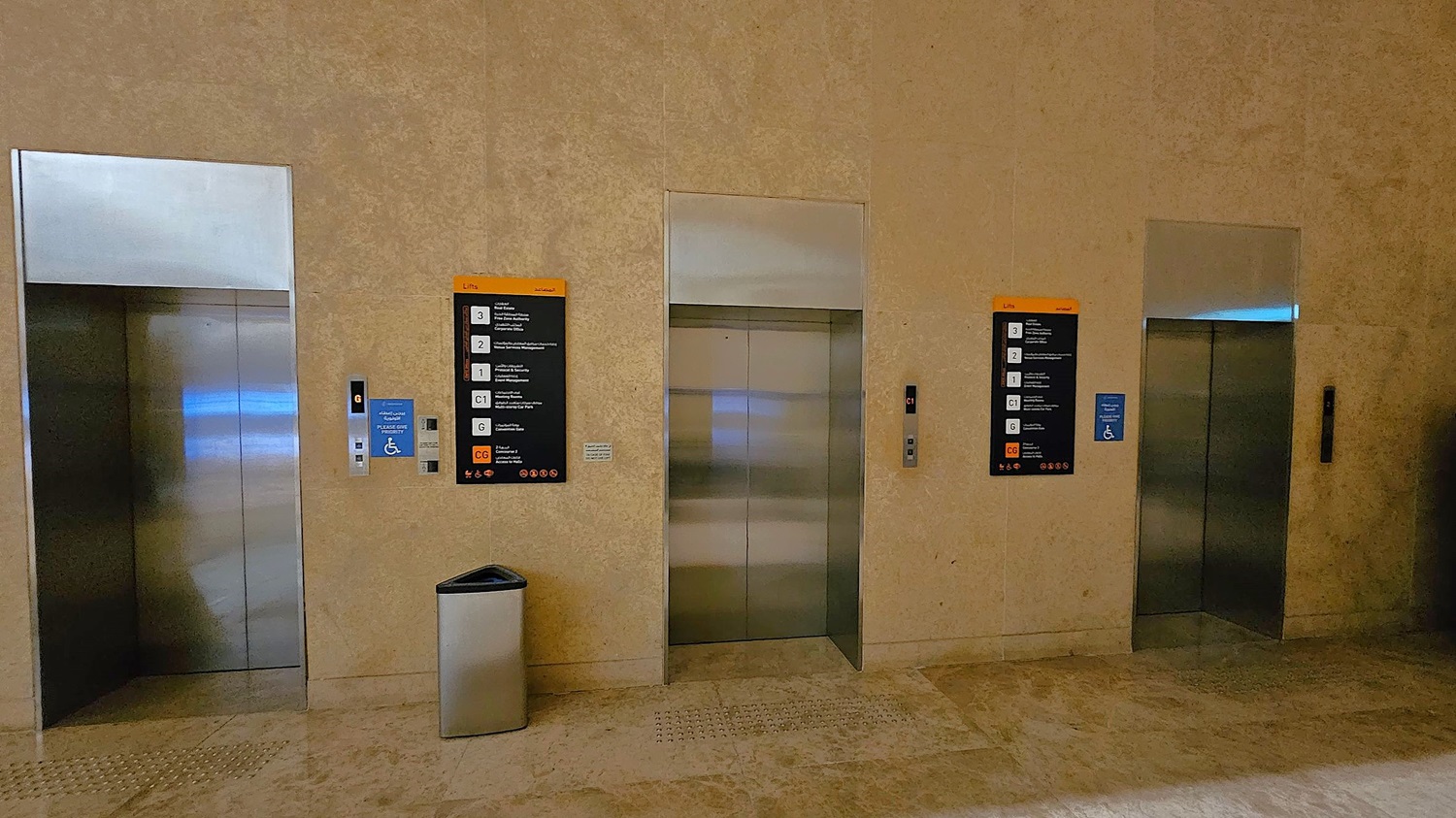 Three lift doors are set into a beige stone wall. Each lift has a metallic finish. On the left, a blue sign indicates accessibility, and a control panel is visible. In the centre, a black and orange directory lists floor numbers and destinations. A silver bin is placed on the floor in front of the middle lift. The floor is made of polished stone tiles. The ceiling is not visible. Three lift doors are set into a beige stone wall. Each lift has a metallic finish. On the left, a blue sign indicates accessibility, and a control panel is visible. In the centre, a black and orange directory lists floor numbers and destinations. A silver bin is placed on the floor in front of the middle lift. The floor is made of polished stone tiles. The ceiling is not visible.