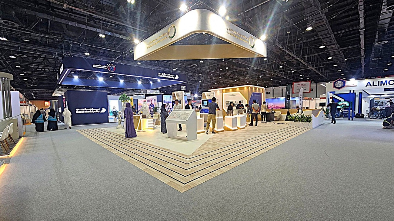 A large exhibition hall with a central booth featuring a wooden floor and illuminated displays. Above, a suspended sign reads "Dubai Municipality." On the left, a group of people walk past a dark display screen. On the right, another booth with the name "ALIMCO" is visible. The ceiling is high with exposed beams and lighting fixtures. The floor is a mix of carpet and wood. A large exhibition hall with a central booth featuring a wooden floor and illuminated displays. Above, a suspended sign reads "Dubai Municipality." On the left, a group of people walk past a dark display screen. On the right, another booth with the name "ALIMCO" is visible. The ceiling is high with exposed beams and lighting fixtures. The floor is a mix of carpet and wood.