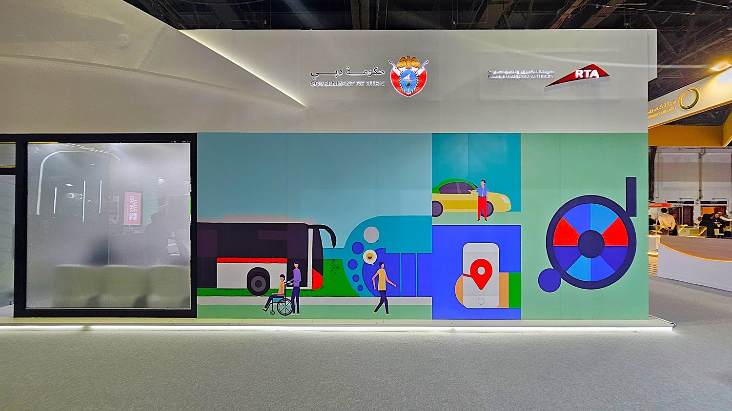 A large wall display with a colourful mural featuring transportation themes. On the left, there's a frosted glass panel with a faint reflection of an interior space. The mural includes illustrations of a bus, a person in a wheelchair, a car, and a smartphone with a location pin. Above, the logos of the Government of Dubai and the Roads & Transport Authority (RTA) are visible. The floor is grey, and the ceiling is dark with lighting fixtures. A large wall display with a colourful mural featuring transportation themes. On the left, there's a frosted glass panel with a faint reflection of an interior space. The mural includes illustrations of a bus, a person in a wheelchair, a car, and a smartphone with a location pin. Above, the logos of the Government of Dubai and the Roads & Transport Authority (RTA) are visible. The floor is grey, and the ceiling is dark with lighting fixtures.