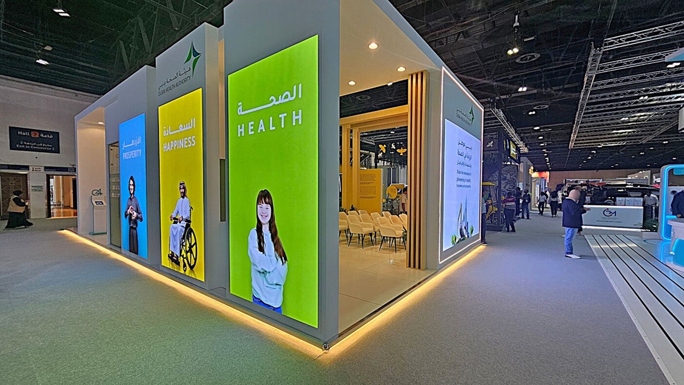 A modern exhibition booth with large vertical screens displaying the words "Prosperity," "Happiness," and "Health" in both English and Arabic. The screens feature images of diverse individuals. Inside the booth, there are rows of white chairs. The floor is carpeted in grey, and the ceiling has exposed beams with spotlights. To the right, people are walking through the exhibition hall, and there are various other booths visible in the background. A modern exhibition booth with large vertical screens displaying the words "Prosperity," "Happiness," and "Health" in both English and Arabic. The screens feature images of diverse individuals. Inside the booth, there are rows of white chairs. The floor is carpeted in grey, and the ceiling has exposed beams with spotlights. To the right, people are walking through the exhibition hall, and there are various other booths visible in the background.