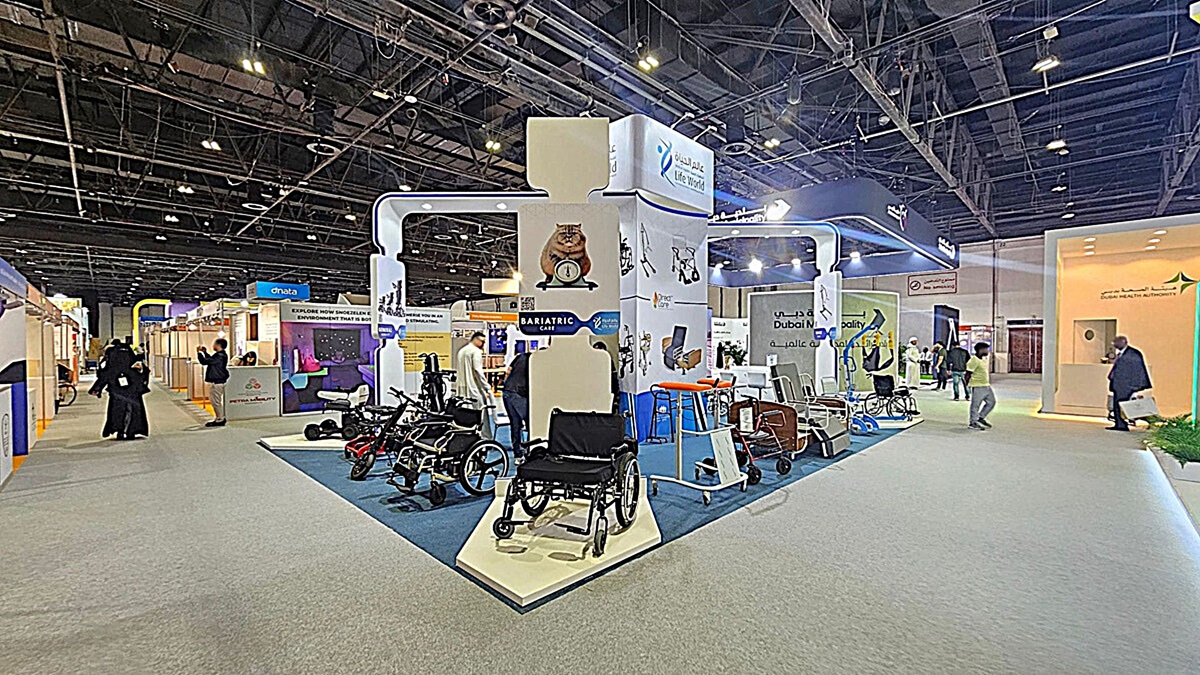 A trade show booth with various mobility aids, including wheelchairs and walkers, displayed on a blue carpeted platform. The booth features signage for bariatric care and is surrounded by other exhibition stands. People are walking around, engaging with the displays. The ceiling is high with exposed beams and lighting fixtures. The floor is covered with grey carpet. A trade show booth with various mobility aids, including wheelchairs and walkers, displayed on a blue carpeted platform. The booth features signage for bariatric care and is surrounded by other exhibition stands. People are walking around, engaging with the displays. The ceiling is high with exposed beams and lighting fixtures. The floor is covered with grey carpet.