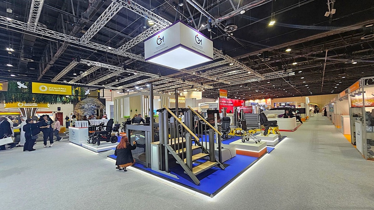 A trade show floor with various exhibits. In the centre, there's a display featuring a small staircase and platform on a blue carpet. Above, a large hanging sign with a logo. To the left, people are gathered near a booth with a green and yellow sign. On the right, more exhibits and a long aisle. The ceiling is high with exposed beams and lighting fixtures. The floor is carpeted in grey. A trade show floor with various exhibits. In the centre, there's a display featuring a small staircase and platform on a blue carpet. Above, a large hanging sign with a logo. To the left, people are gathered near a booth with a green and yellow sign. On the right, more exhibits and a long aisle. The ceiling is high with exposed beams and lighting fixtures. The floor is carpeted in grey.