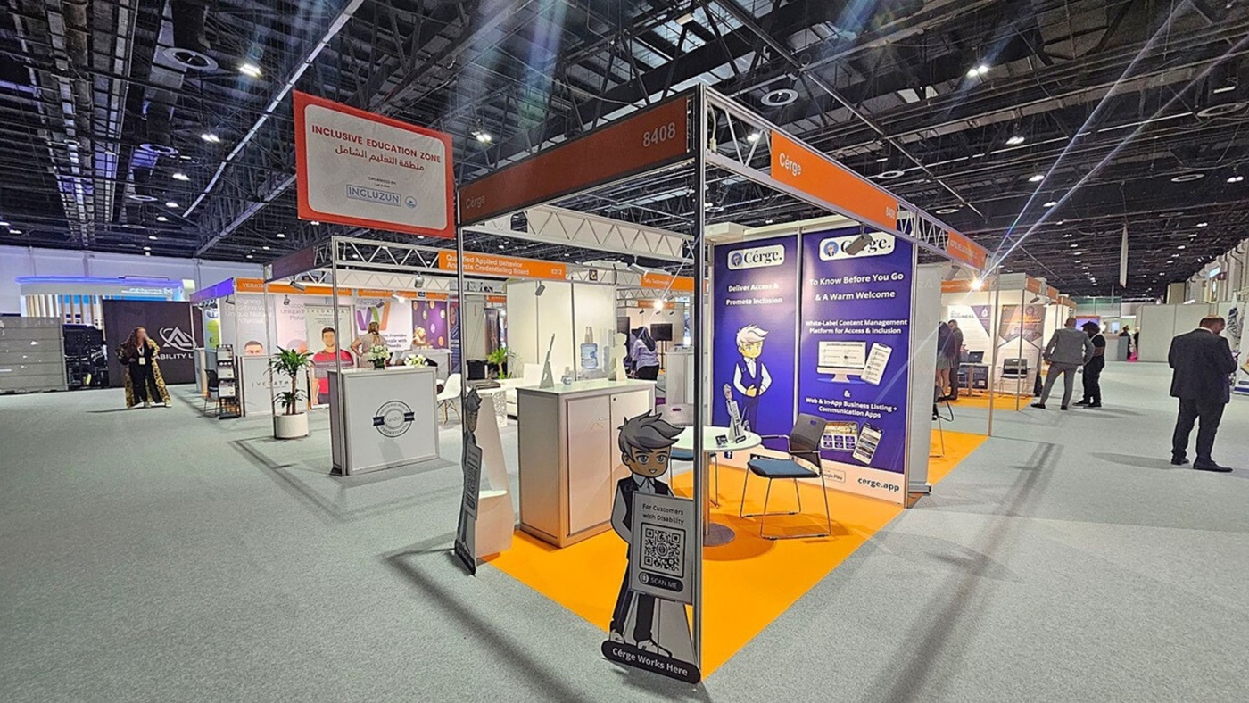 A trade show booth with a sign reading "Inclusive Education Zone" in both English and Arabic. The booth features a display for "Cérge," with promotional materials and a cartoon character holding a QR code sign. The floor is carpeted in grey, and the booth area is marked with an orange carpet. Several people are walking around the exhibition hall, which has a high ceiling with exposed beams and lighting fixtures. Other booths are visible in the background. A trade show booth with a sign reading "Inclusive Education Zone" in both English and Arabic. The booth features a display for "Cérge," with promotional materials and a cartoon character holding a QR code sign. The floor is carpeted in grey, and the booth area is marked with an orange carpet. Several people are walking around the exhibition hall, which has a high ceiling with exposed beams and lighting fixtures. Other booths are visible in the background.