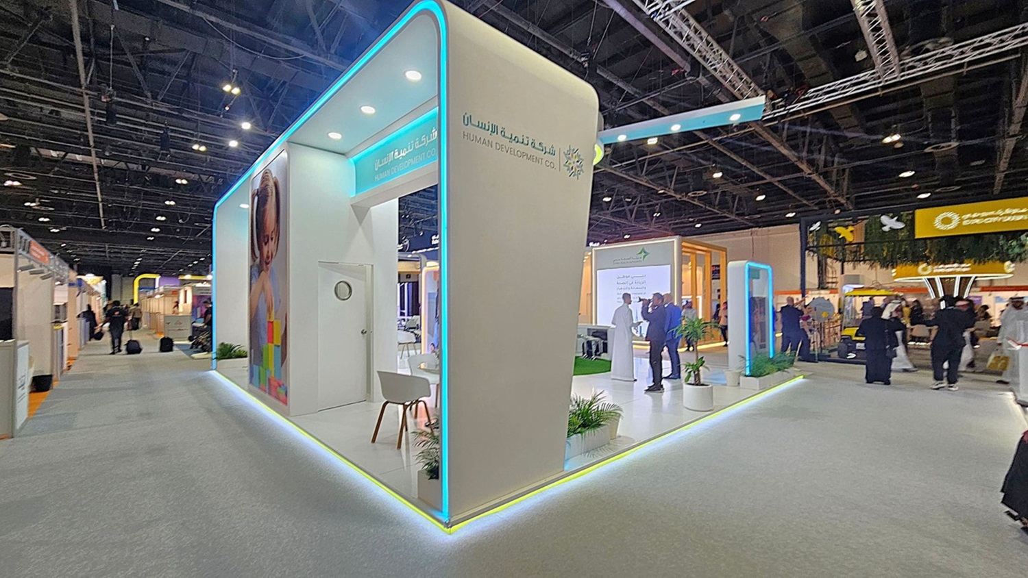 A trade show booth with a modern design featuring a white structure with blue neon lighting accents. On the left, there's a large image of a child and a small seating area with a white chair. The booth is branded with "Human Development Co." in both English and Arabic. In the background, people are walking around, and other booths are visible, including one with a yellow sign. The ceiling is high with exposed beams and lighting fixtures. The floor is carpeted in grey. A trade show booth with a modern design featuring a white structure with blue neon lighting accents. On the left, there's a large image of a child and a small seating area with a white chair. The booth is branded with "Human Development Co." in both English and Arabic. In the background, people are walking around, and other booths are visible, including one with a yellow sign. The ceiling is high with exposed beams and lighting fixtures. The floor is carpeted in grey.