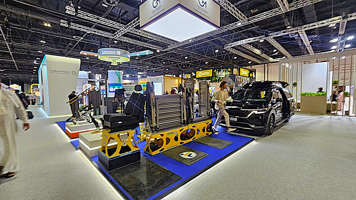 A trade show floor with various exhibits. On the left, there are display stands with equipment and a person walking by. In the centre, a platform with mechanical components and seating is showcased on a blue mat. On the right, a black vehicle with open doors is displayed. The background features booths with signage and people interacting. The ceiling has a grid of lights and metal trusses. The floor is carpeted in grey. A trade show floor with various exhibits. On the left, there are display stands with equipment and a person walking by. In the centre, a platform with mechanical components and seating is showcased on a blue mat. On the right, a black vehicle with open doors is displayed. The background features booths with signage and people interacting. The ceiling has a grid of lights and metal trusses. The floor is carpeted in grey.