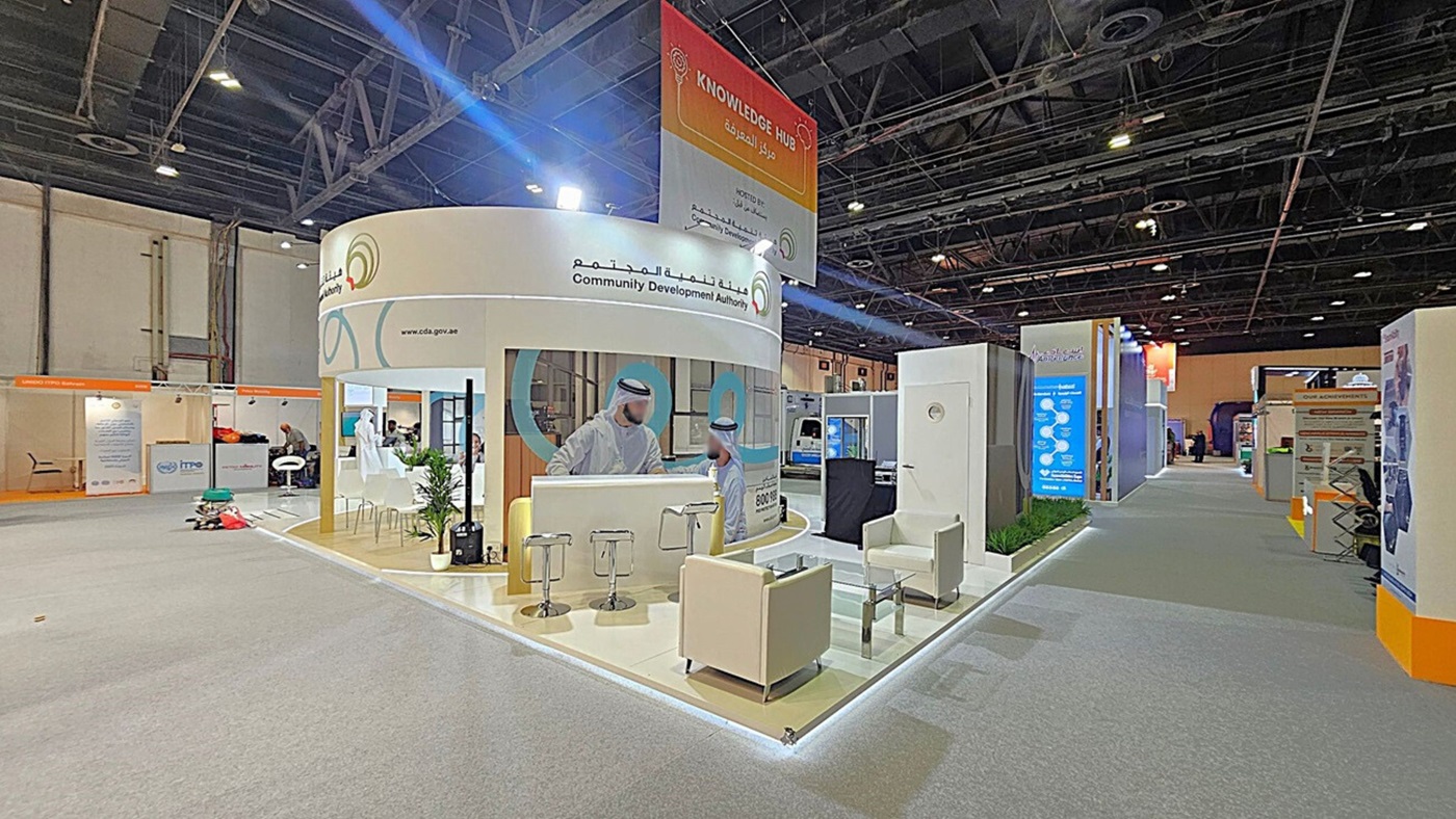 A trade show booth with a circular design, featuring the Community Development Authority. The booth includes a counter with stools, a glass table, and white armchairs. A large poster with two men in traditional attire is displayed. The floor is carpeted in grey, and the ceiling has exposed beams and lighting. Adjacent booths and informational displays are visible along the aisle. A "Knowledge Hub" sign hangs above. A trade show booth with a circular design, featuring the Community Development Authority. The booth includes a counter with stools, a glass table, and white armchairs. A large poster with two men in traditional attire is displayed. The floor is carpeted in grey, and the ceiling has exposed beams and lighting. Adjacent booths and informational displays are visible along the aisle. A "Knowledge Hub" sign hangs above.