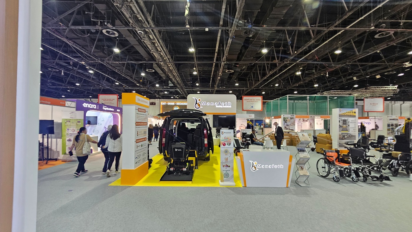 A trade show booth with a black vehicle in the centre, featuring an accessibility ramp extending onto a bright yellow platform. The booth is branded with "Manafeth" and includes informational displays. On the right, there are various mobility aids like wheelchairs. In the background, other booths are visible with people walking around. The ceiling is industrial with exposed beams and lighting fixtures. The floor is carpeted in grey. A trade show booth with a black vehicle in the centre, featuring an accessibility ramp extending onto a bright yellow platform. The booth is branded with "Manafeth" and includes informational displays. On the right, there are various mobility aids like wheelchairs. In the background, other booths are visible with people walking around. The ceiling is industrial with exposed beams and lighting fixtures. The floor is carpeted in grey.