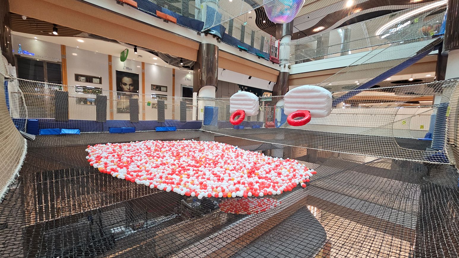 A large indoor play area with a netted floor filled with red and white plastic balls in the centre. Two inflatable white structures with red rings are suspended above the net. The background features a multi-level shopping centre with glass railings, columns, and various shopfronts. The ceiling has modern lighting fixtures, and the floor beneath the net is visible through the mesh. A large indoor play area with a netted floor filled with red and white plastic balls in the centre. Two inflatable white structures with red rings are suspended above the net. The background features a multi-level shopping centre with glass railings, columns, and various shopfronts. The ceiling has modern lighting fixtures, and the floor beneath the net is visible through the mesh.