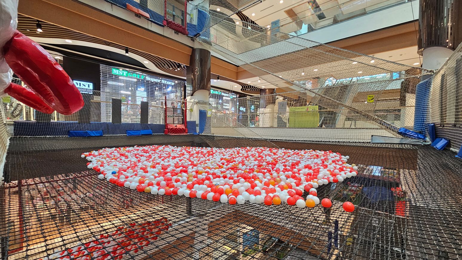 A shopping centre interior with a large netted play area in the foreground, filled with red, white, and orange plastic balls. On the left, a large red inflatable object is visible. The background features a store with a sign reading "MST GOLF Super Store" and other shops. The ceiling is high with multiple levels and glass railings, and the floor is covered with netting. The area is well-lit with overhead lights. A shopping centre interior with a large netted play area in the foreground, filled with red, white, and orange plastic balls. On the left, a large red inflatable object is visible. The background features a store with a sign reading "MST GOLF Super Store" and other shops. The ceiling is high with multiple levels and glass railings, and the floor is covered with netting. The area is well-lit with overhead lights.