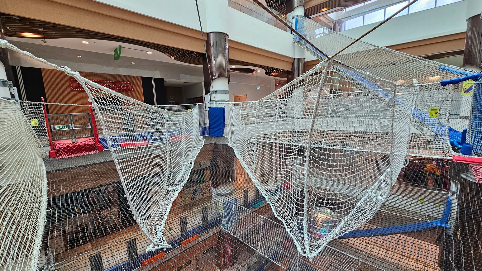 A large indoor play area with suspended net walkways and platforms. The nets are white and form a pathway across multiple levels. In the background, there are wooden panels and a red exit sign. The area is supported by large cylindrical columns. The floor below is visible through the nets, showing a lower level with various activities. The ceiling has a modern design with recessed lighting and skylights. A large indoor play area with suspended net walkways and platforms. The nets are white and form a pathway across multiple levels. In the background, there are wooden panels and a red exit sign. The area is supported by large cylindrical columns. The floor below is visible through the nets, showing a lower level with various activities. The ceiling has a modern design with recessed lighting and skylights.