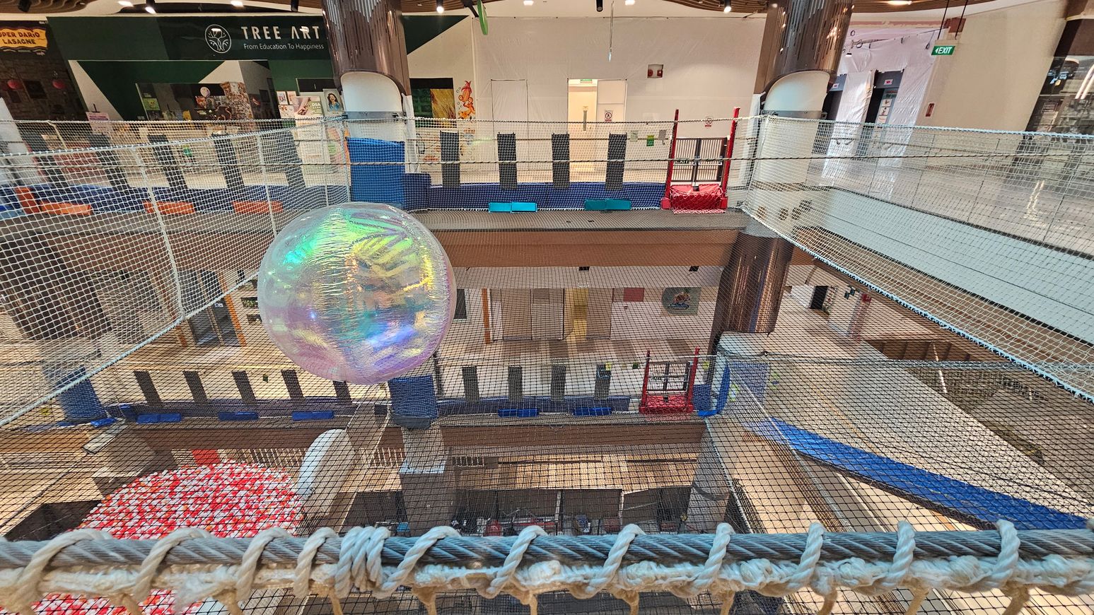 A large, transparent, iridescent ball is suspended within a netted play area. The netting is extensive, covering multiple levels of a shopping centre. In the background, there is a shop with a sign reading "Tree Art" and various artworks displayed. The floor below is visible through the netting, showing a mix of open spaces and other structures. The area is brightly lit with overhead lights, and there are red and blue play structures on the right side. A large, transparent, iridescent ball is suspended within a netted play area. The netting is extensive, covering multiple levels of a shopping centre. In the background, there is a shop with a sign reading "Tree Art" and various artworks displayed. The floor below is visible through the netting, showing a mix of open spaces and other structures. The area is brightly lit with overhead lights, and there are red and blue play structures on the right side.