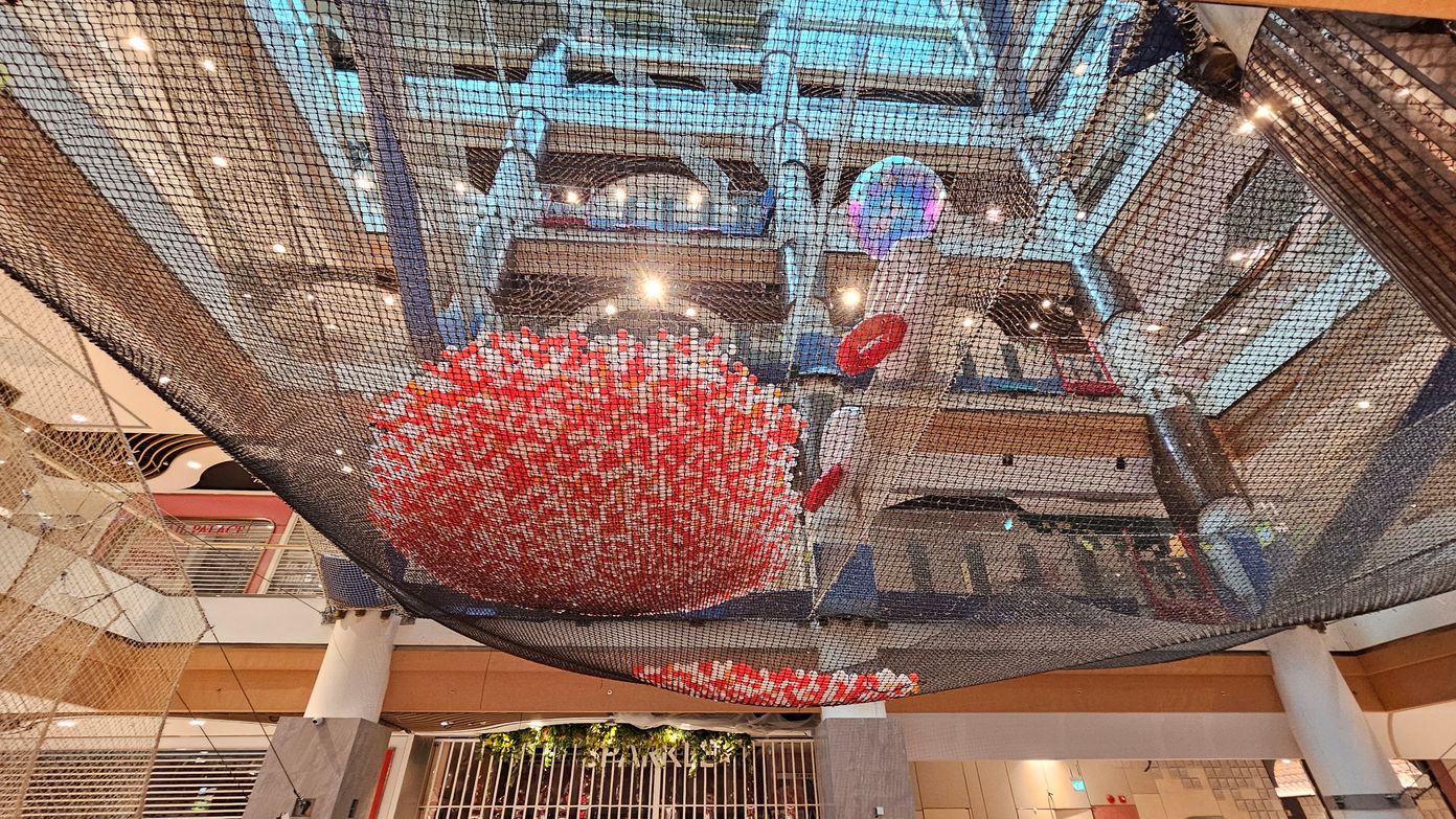 A large indoor space with a net structure suspended from the ceiling. In the centre, there's a spherical installation made of red and white elements. The background shows multiple floors with railings and a variety of lights. The ceiling is high with visible beams and spotlights. The floor is not visible, but there are columns supporting the structure. Some greenery is visible on the lower level. A large indoor space with a net structure suspended from the ceiling. In the centre, there's a spherical installation made of red and white elements. The background shows multiple floors with railings and a variety of lights. The ceiling is high with visible beams and spotlights. The floor is not visible, but there are columns supporting the structure. Some greenery is visible on the lower level.