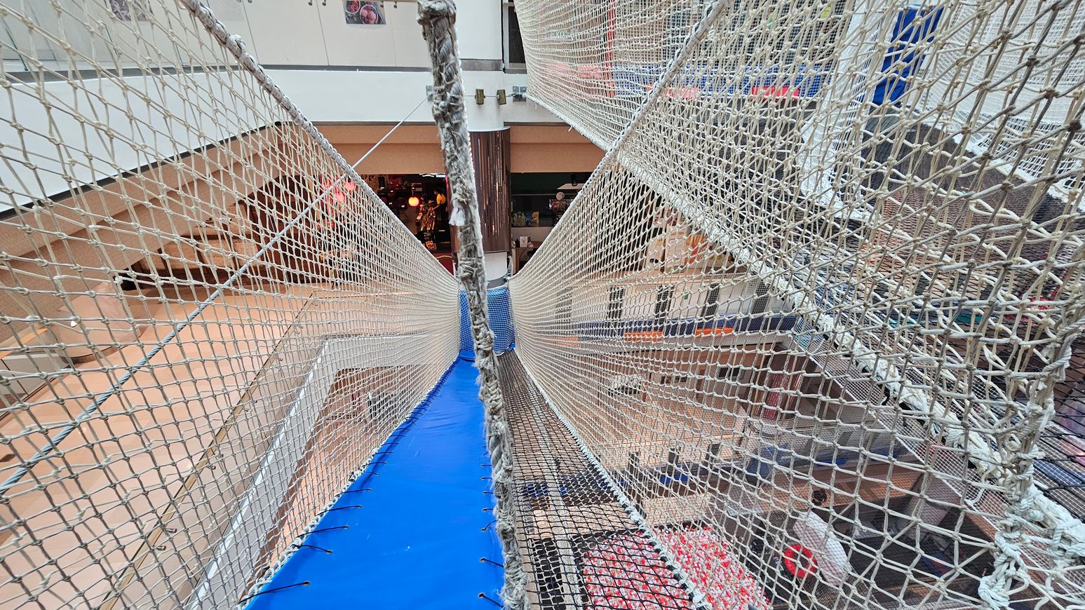 A suspended net bridge with blue padding on the floor stretches across a multi-level indoor space. The netting is tightly woven, forming walls on either side. Below, various floors of a shopping centre are visible, with shops and people. The background shows a glass railing and a column, with some lights and displays from the shops. The ceiling is not visible. A suspended net bridge with blue padding on the floor stretches across a multi-level indoor space. The netting is tightly woven, forming walls on either side. Below, various floors of a shopping centre are visible, with shops and people. The background shows a glass railing and a column, with some lights and displays from the shops. The ceiling is not visible.