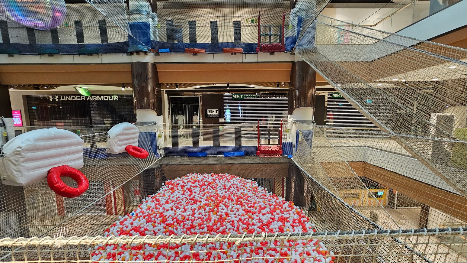 A shopping centre with a netted play area suspended above the ground. In the foreground, there are large white inflatable structures with red rings attached. Below, a pit filled with red and white balls. The background shows shopfronts, including Under Armour and MST, with closed shutters. The area is enclosed with netting, and there are multiple levels visible, with glass railings and a mix of wooden and metal elements. A shopping centre with a netted play area suspended above the ground. In the foreground, there are large white inflatable structures with red rings attached. Below, a pit filled with red and white balls. The background shows shopfronts, including Under Armour and MST, with closed shutters. The area is enclosed with netting, and there are multiple levels visible, with glass railings and a mix of wooden and metal elements.