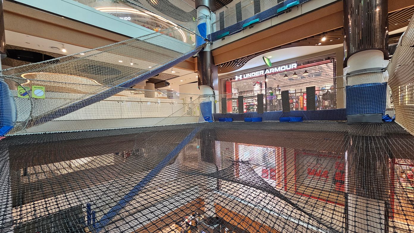 A shopping centre interior with multiple levels. In the foreground, there is a large net structure spanning across the space, supported by blue and black padding. On the right, a shop with a visible "Under Armour" sign. The background shows another level with a curved ceiling design and various lights. The floor is tiled, and there are signs indicating "No Entry" on the netting. A shopping centre interior with multiple levels. In the foreground, there is a large net structure spanning across the space, supported by blue and black padding. On the right, a shop with a visible "Under Armour" sign. The background shows another level with a curved ceiling design and various lights. The floor is tiled, and there are signs indicating "No Entry" on the netting.