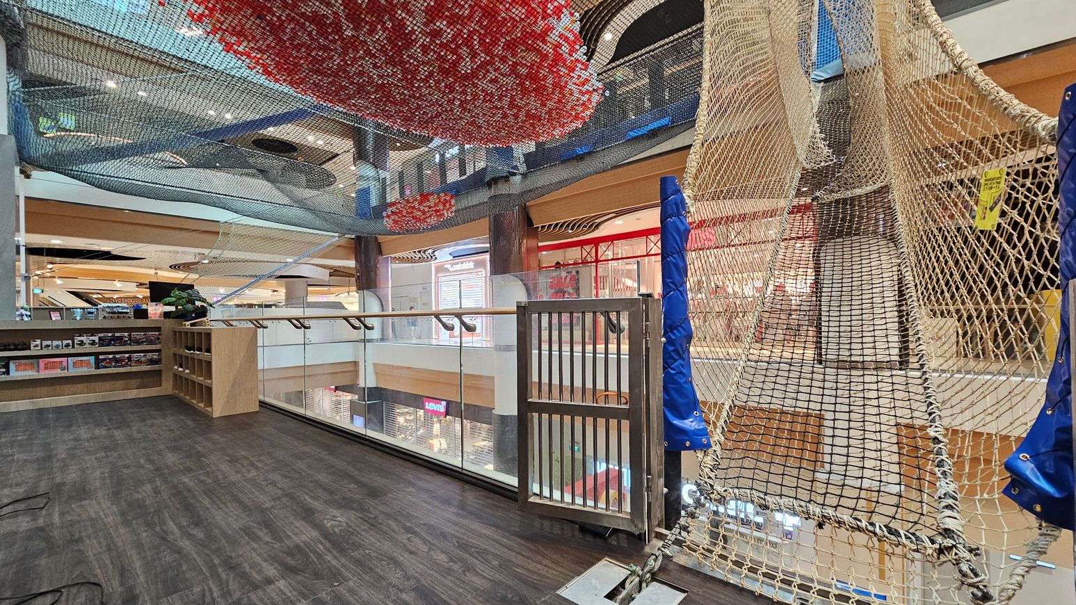A shopping centre interior with a dark wooden floor. On the right, a netted play area with blue padding is suspended, leading upwards. In the middle, a glass railing overlooks the lower level, where shop signs like Levi's are visible. On the left, wooden shelves display various items. Above, colourful netting in red and blue is suspended from the ceiling, which features spotlights and wooden beams. A shopping centre interior with a dark wooden floor. On the right, a netted play area with blue padding is suspended, leading upwards. In the middle, a glass railing overlooks the lower level, where shop signs like Levi's are visible. On the left, wooden shelves display various items. Above, colourful netting in red and blue is suspended from the ceiling, which features spotlights and wooden beams.