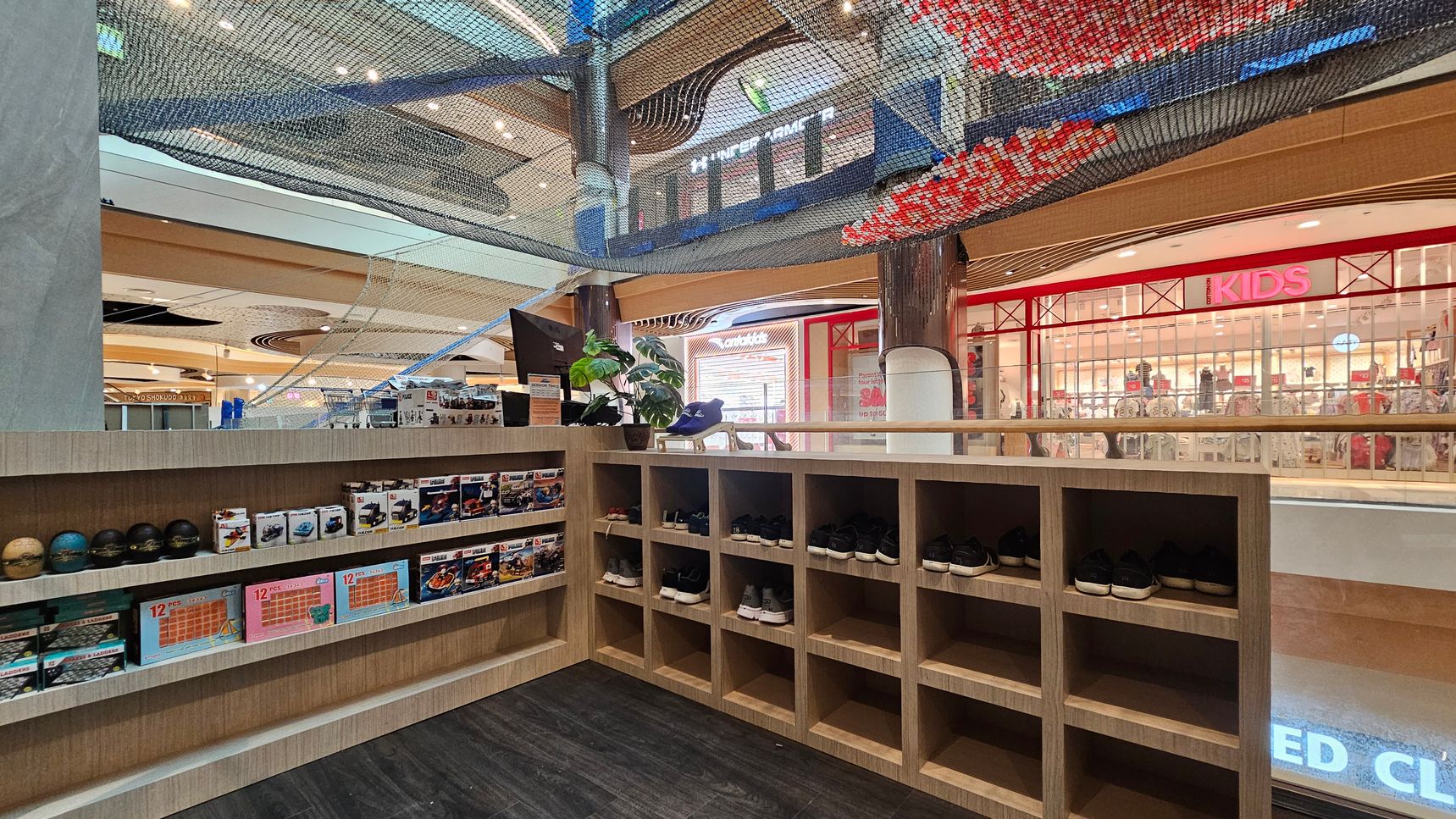 On the left, there are shelves displaying various boxed toys and small balls. In the middle, a wooden counter holds a plant and a pair of shoes. On the right, there are cubbyholes with more shoes. The background features a shop with a red sign reading "KIDS" and a glass storefront. Above, a netted structure is visible, and the ceiling has circular light fixtures. The floor is dark and smooth. On the left, there are shelves displaying various boxed toys and small balls. In the middle, a wooden counter holds a plant and a pair of shoes. On the right, there are cubbyholes with more shoes. The background features a shop with a red sign reading "KIDS" and a glass storefront. Above, a netted structure is visible, and the ceiling has circular light fixtures. The floor is dark and smooth.