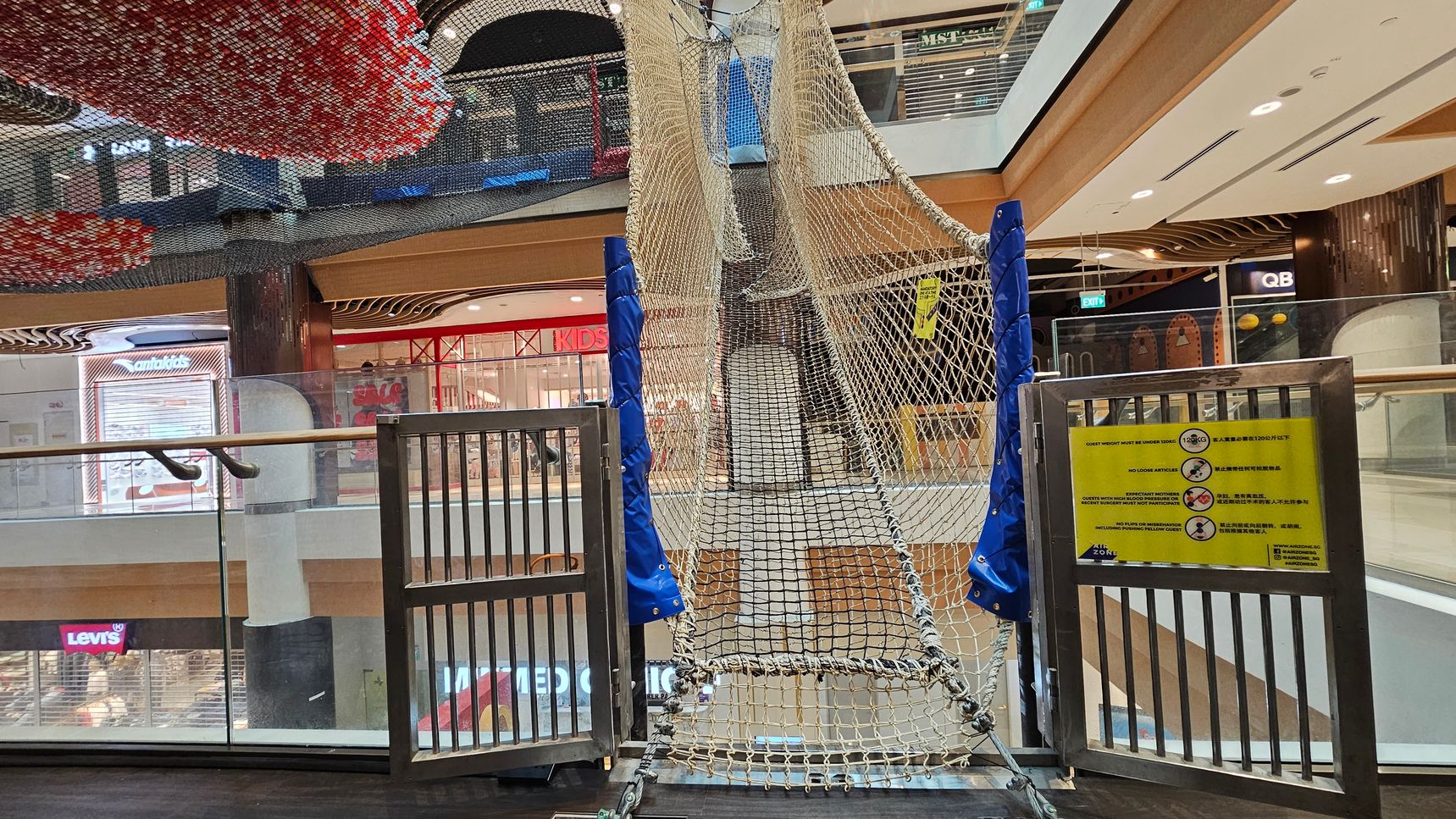 A shopping centre interior with a netted climbing structure in the foreground, supported by blue padded poles. A metal gate with a yellow sign displaying safety instructions is attached to the structure. In the background, there are various shopfronts, including Levi's and a children's store. The ceiling features decorative elements with red and black patterns. The floor is dark, and glass railings are visible on the left side. A shopping centre interior with a netted climbing structure in the foreground, supported by blue padded poles. A metal gate with a yellow sign displaying safety instructions is attached to the structure. In the background, there are various shopfronts, including Levi's and a children's store. The ceiling features decorative elements with red and black patterns. The floor is dark, and glass railings are visible on the left side.
