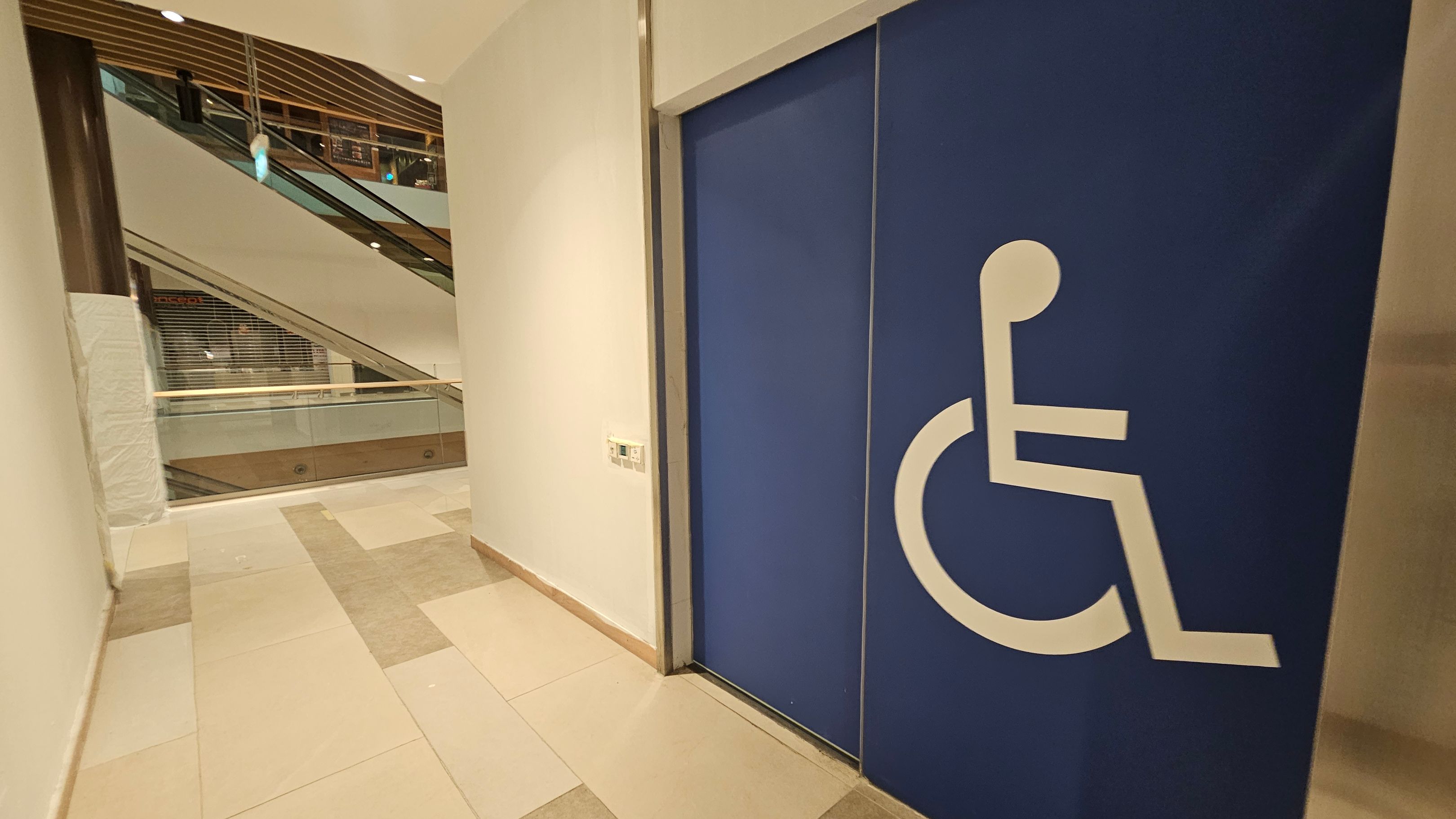 On the right, there is a large blue door with a white wheelchair symbol, indicating accessibility. The left side shows a corridor with a view of an escalator and glass railings. The floor is tiled in shades of beige and pink. The ceiling has recessed lighting and wooden slats. On the right, there is a large blue door with a white wheelchair symbol, indicating accessibility. The left side shows a corridor with a view of an escalator and glass railings. The floor is tiled in shades of beige and pink. The ceiling has recessed lighting and wooden slats.
