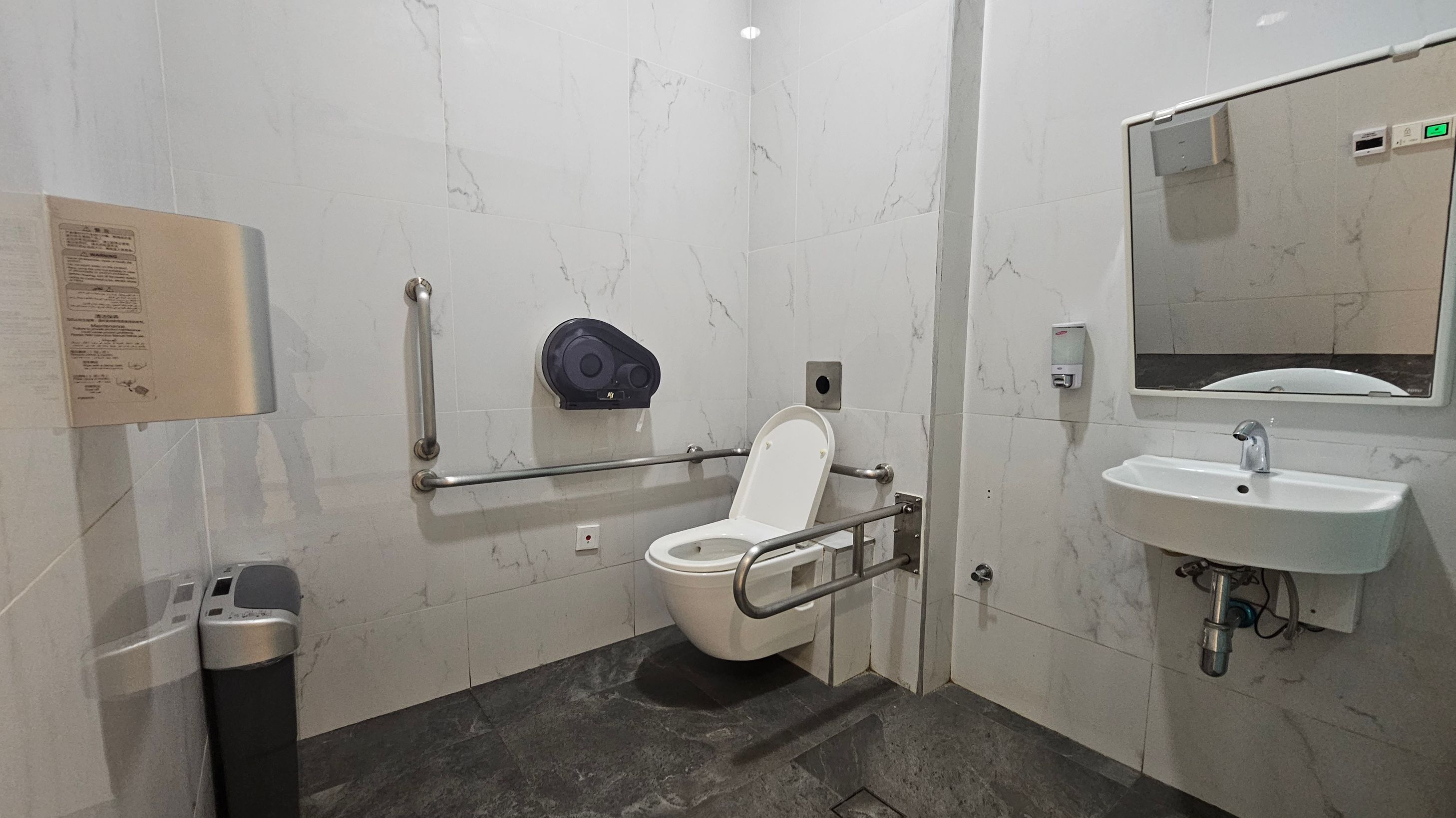 A public restroom with a white toilet featuring a raised seat and support rails on both sides. To the left, a waste bin and a wall-mounted instruction panel are visible. On the right, a white sink with a mirror above it and a soap dispenser to the side. The walls are covered in light-coloured tiles with a subtle marble pattern, and the floor is dark grey. A public restroom with a white toilet featuring a raised seat and support rails on both sides. To the left, a waste bin and a wall-mounted instruction panel are visible. On the right, a white sink with a mirror above it and a soap dispenser to the side. The walls are covered in light-coloured tiles with a subtle marble pattern, and the floor is dark grey.