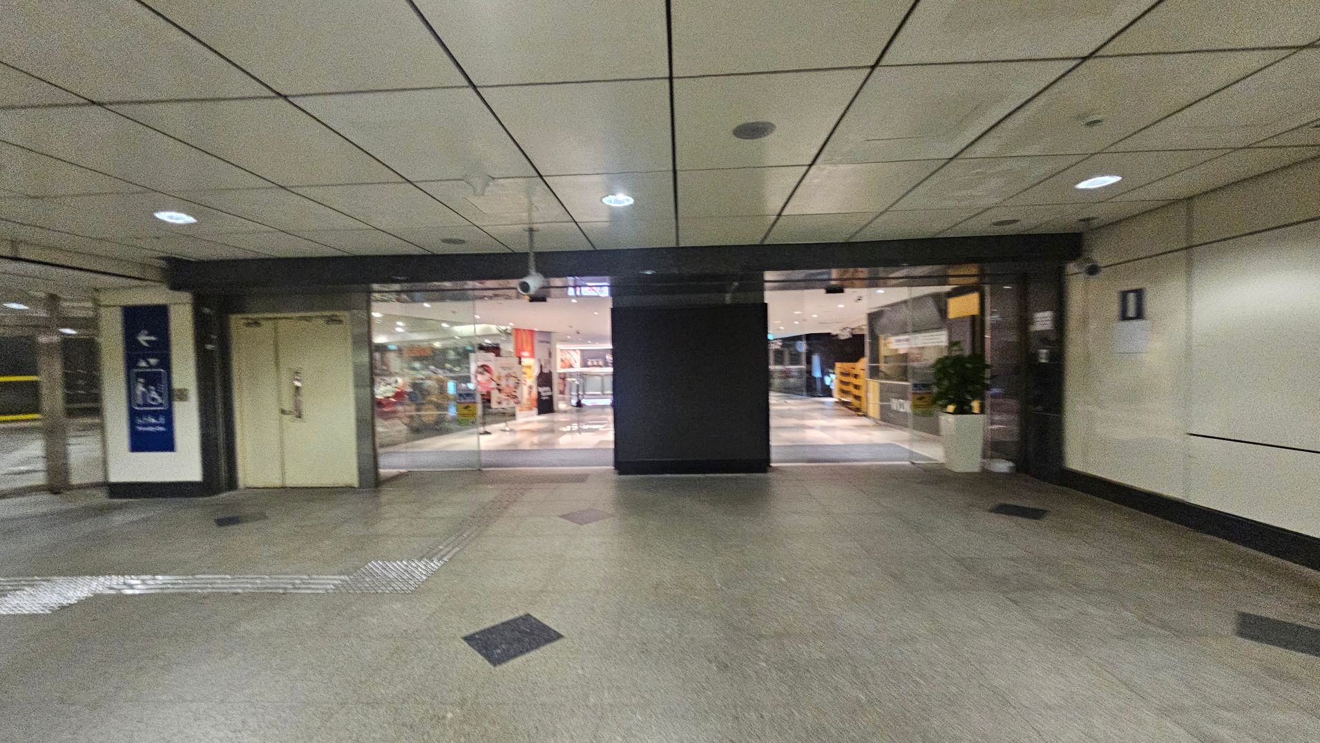 A corridor with a tiled ceiling and greenish floor. In the centre, there are glass doors leading to a brightly lit interior space with visible signage and displays. On the left, there's a sign with directional arrows and a closed door. On the right, a wall with a small sign and a potted plant near the entrance. The area is well-lit with ceiling lights. A corridor with a tiled ceiling and greenish floor. In the centre, there are glass doors leading to a brightly lit interior space with visible signage and displays. On the left, there's a sign with directional arrows and a closed door. On the right, a wall with a small sign and a potted plant near the entrance. The area is well-lit with ceiling lights.