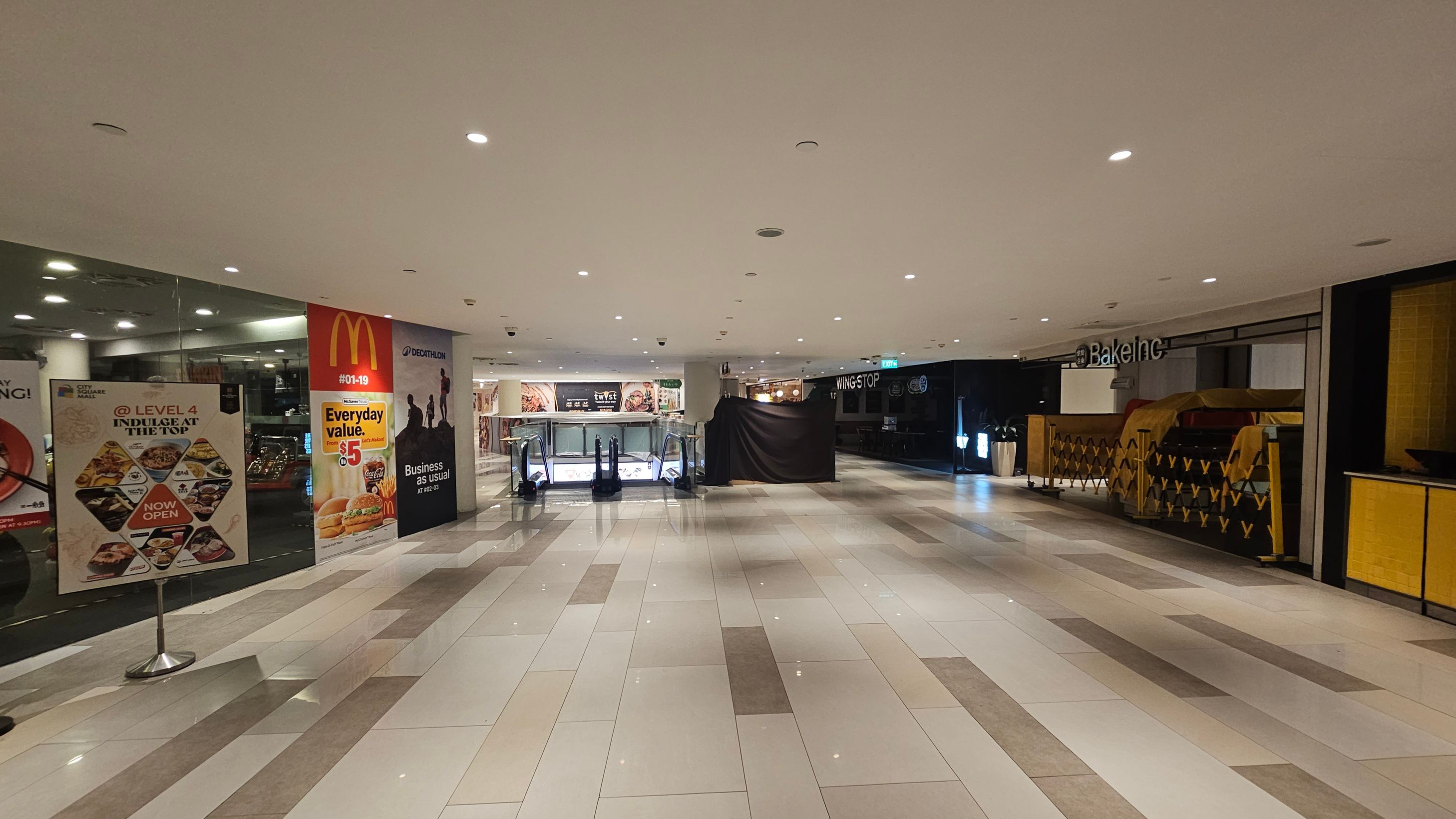 A shopping mall interior with a wide, tiled floor in shades of grey and white. On the left, there are promotional posters, including one for McDonald's. In the centre, an escalator is visible, leading to a lower level. On the right, there is a closed shop with a sign reading "Bake Inc" and a covered counter. The ceiling is white with recessed lighting, creating a bright atmosphere. A shopping mall interior with a wide, tiled floor in shades of grey and white. On the left, there are promotional posters, including one for McDonald's. In the centre, an escalator is visible, leading to a lower level. On the right, there is a closed shop with a sign reading "Bake Inc" and a covered counter. The ceiling is white with recessed lighting, creating a bright atmosphere.