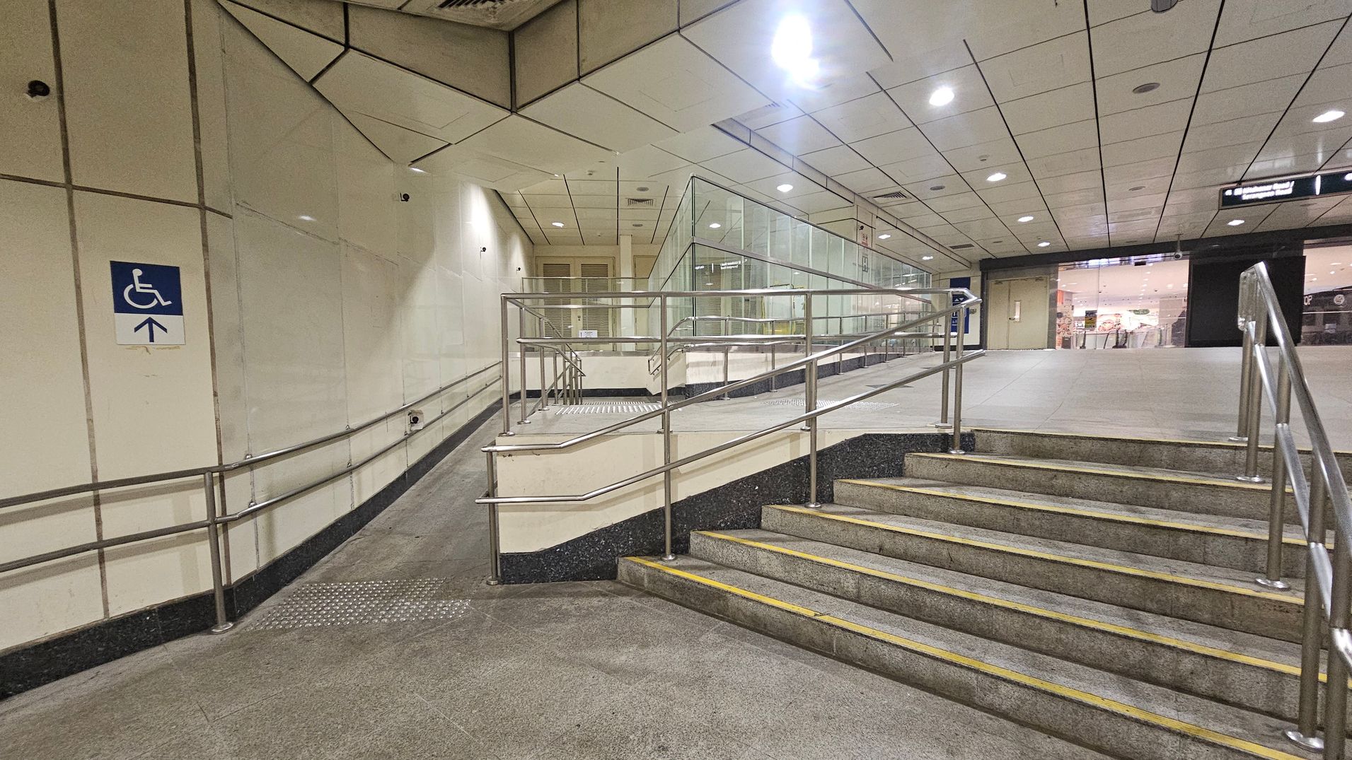 A public indoor space with a ramp and stairs. On the left, a ramp with metal railings leads upwards, marked by a blue sign indicating accessibility. The right side features a set of stairs with similar metal railings. The background shows a glass partition and a view into a well-lit area with shops. The ceiling is tiled with recessed lighting, and the floor is a mix of green and grey tiles. A public indoor space with a ramp and stairs. On the left, a ramp with metal railings leads upwards, marked by a blue sign indicating accessibility. The right side features a set of stairs with similar metal railings. The background shows a glass partition and a view into a well-lit area with shops. The ceiling is tiled with recessed lighting, and the floor is a mix of green and grey tiles.