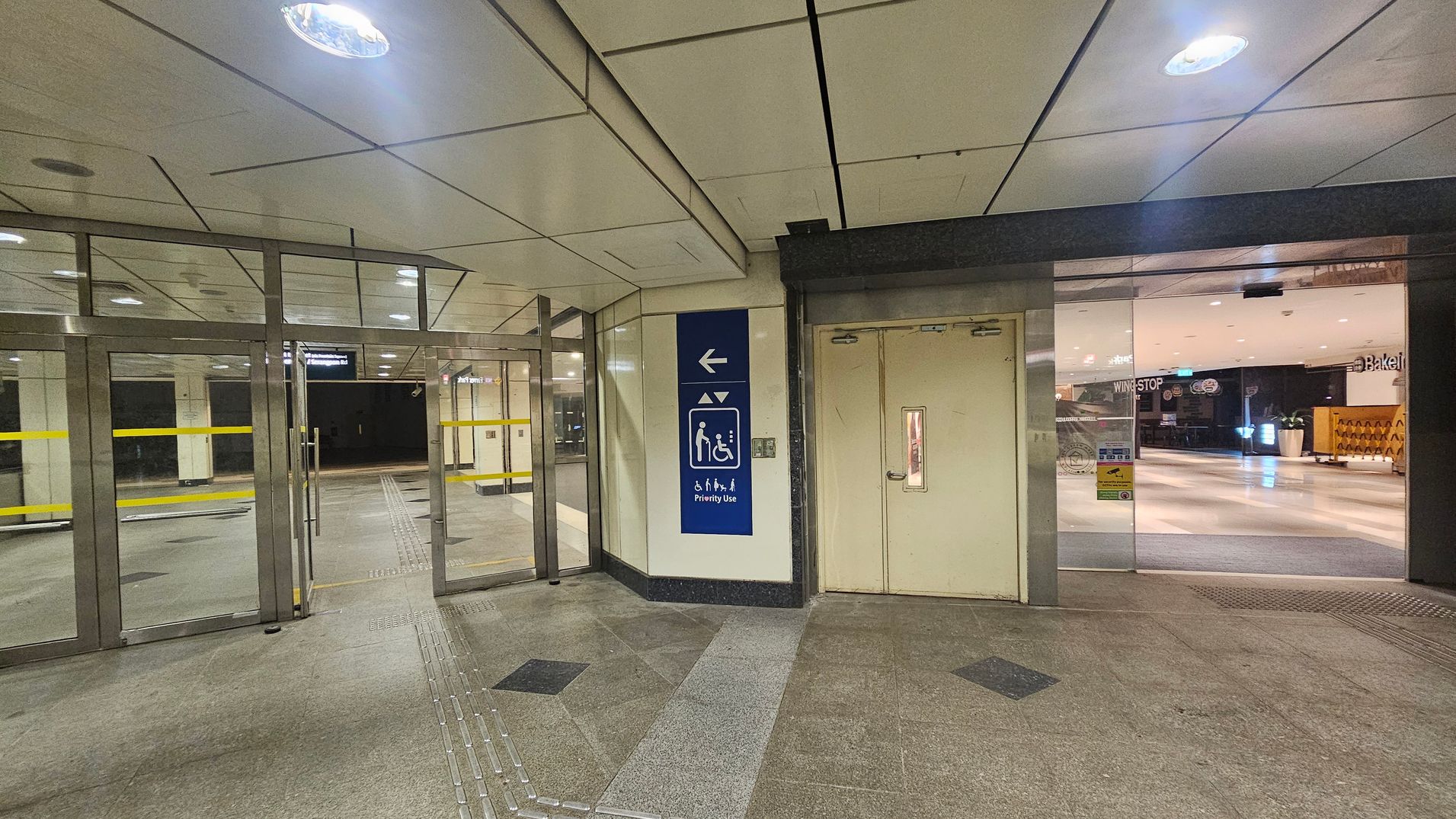 On the left, there are glass doors with yellow horizontal lines, leading to a corridor. In the centre, a blue sign indicates priority use for lifts, with symbols for accessibility. On the right, a closed door with a small window is adjacent to a glass entrance showing a glimpse of a brightly lit interior with shops. The floor is tiled with a mix of grey and black patterns, and the ceiling is white with recessed lighting. On the left, there are glass doors with yellow horizontal lines, leading to a corridor. In the centre, a blue sign indicates priority use for lifts, with symbols for accessibility. On the right, a closed door with a small window is adjacent to a glass entrance showing a glimpse of a brightly lit interior with shops. The floor is tiled with a mix of grey and black patterns, and the ceiling is white with recessed lighting.
