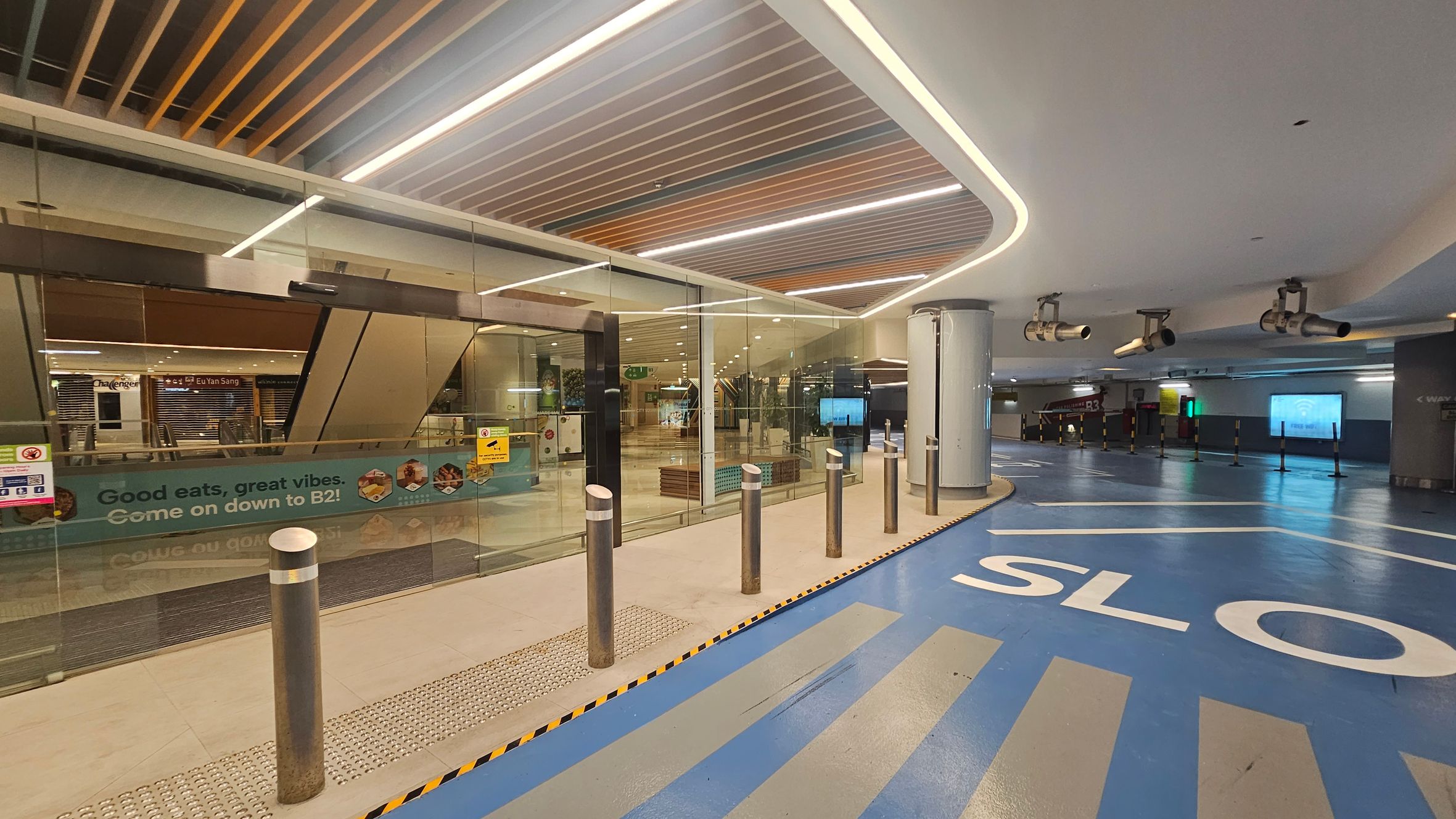 On the left, there is a glass entrance with a sign that reads "Good eats, great vibes. Come on down to B2!" Behind the glass, there are closed shopfronts, including one labelled "Eu Yan Sang." The floor is a mix of beige and blue, with the word "SLOW" painted in white. Metal bollards line the entrance. The ceiling features a modern design with recessed lighting and wooden slats. On the right, there are large cylindrical structures and overhead lights. On the left, there is a glass entrance with a sign that reads "Good eats, great vibes. Come on down to B2!" Behind the glass, there are closed shopfronts, including one labelled "Eu Yan Sang." The floor is a mix of beige and blue, with the word "SLOW" painted in white. Metal bollards line the entrance. The ceiling features a modern design with recessed lighting and wooden slats. On the right, there are large cylindrical structures and overhead lights.