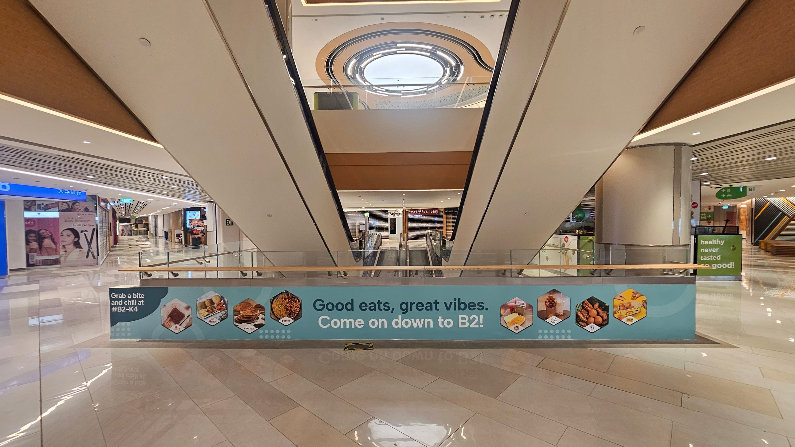 A shopping mall interior with a central escalator leading downwards. A blue banner across the escalator reads, "Good eats, great vibes. Come on down to B2!" with images of food items. The left side has shopfronts with advertisements, while the right side features a pillar and a sign saying "healthy never tasted so good!" The floor is shiny and tiled, and the ceiling has recessed lighting with a circular skylight above. A shopping mall interior with a central escalator leading downwards. A blue banner across the escalator reads, "Good eats, great vibes. Come on down to B2!" with images of food items. The left side has shopfronts with advertisements, while the right side features a pillar and a sign saying "healthy never tasted so good!" The floor is shiny and tiled, and the ceiling has recessed lighting with a circular skylight above.