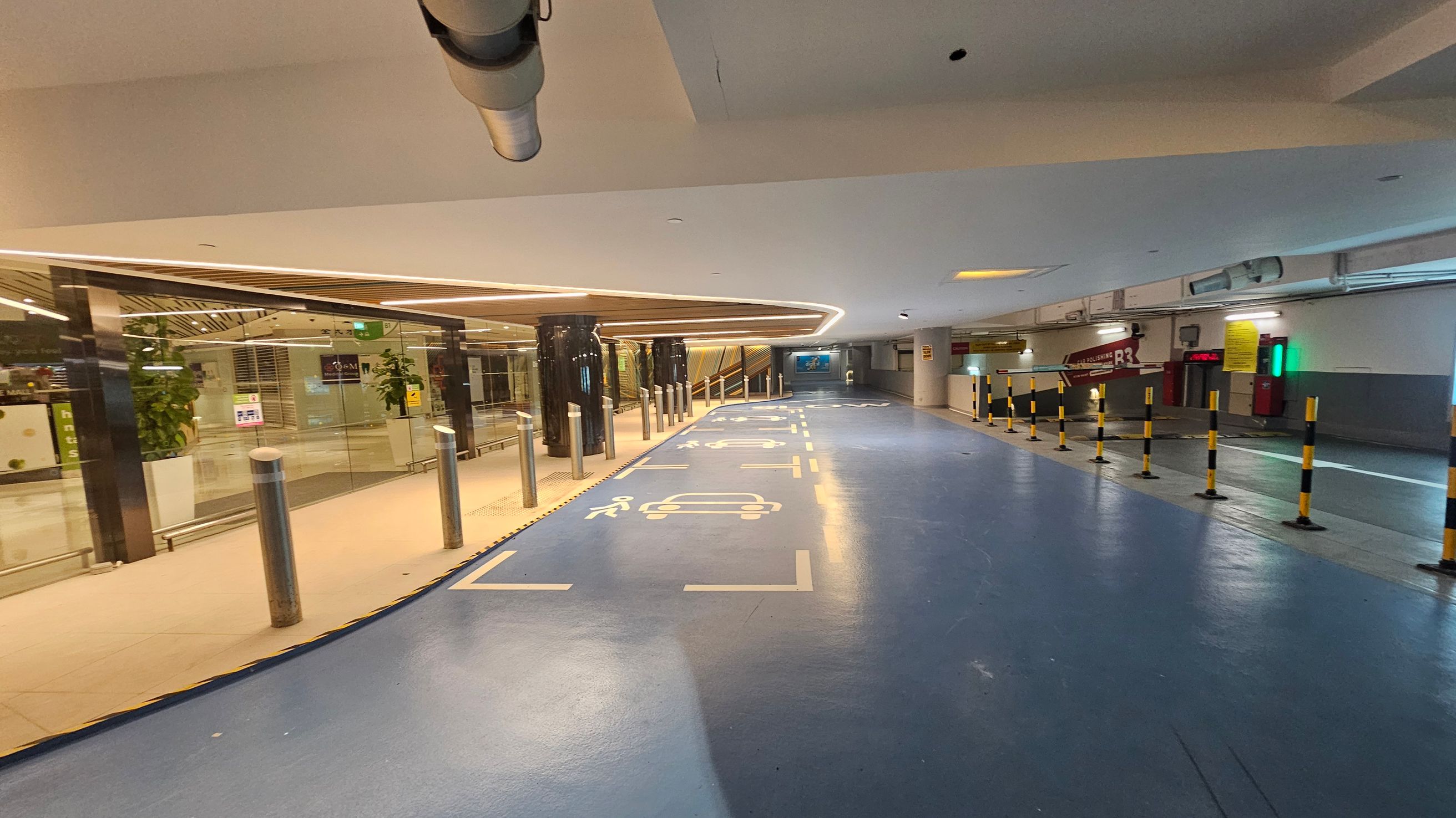 A parking area with a blue floor marked with white symbols for cars and pedestrians. On the left, there is a glass wall with plants and a view into a building interior. The ceiling has recessed lighting and a visible duct. On the right, there are yellow and black striped bollards and a sign indicating "R3". The background shows an escalator and more signage. A parking area with a blue floor marked with white symbols for cars and pedestrians. On the left, there is a glass wall with plants and a view into a building interior. The ceiling has recessed lighting and a visible duct. On the right, there are yellow and black striped bollards and a sign indicating "R3". The background shows an escalator and more signage.