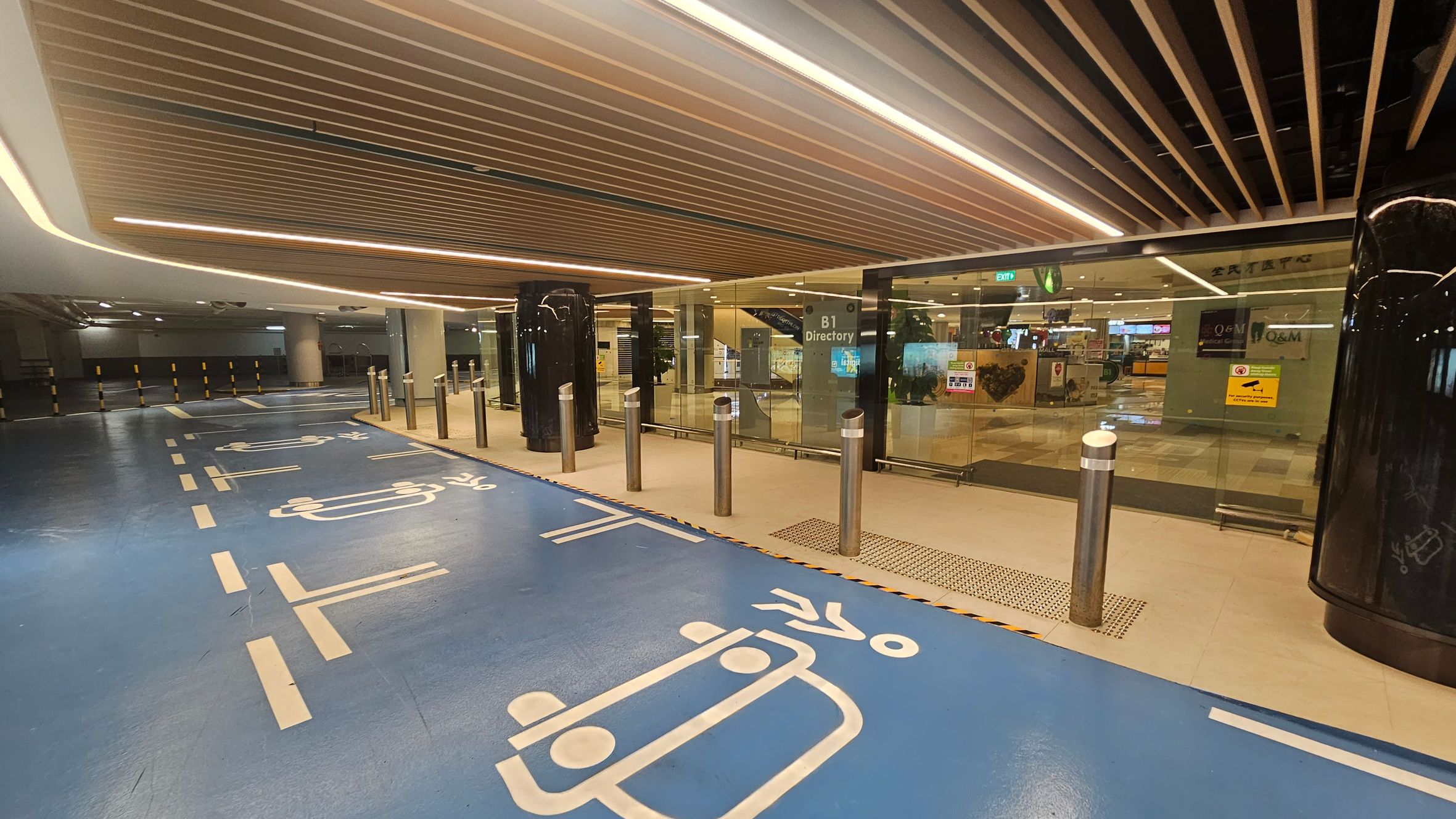 A modern underground parking area with a blue floor featuring white painted symbols for vehicles and pedestrians. On the right, there is a glass entrance with signs, including "B1 Directory," and several bollards in front. The ceiling has a wooden slat design with integrated lighting. In the background, there are visible shops and an exit sign. The left side shows more of the parking area with yellow and black striped barriers. A modern underground parking area with a blue floor featuring white painted symbols for vehicles and pedestrians. On the right, there is a glass entrance with signs, including "B1 Directory," and several bollards in front. The ceiling has a wooden slat design with integrated lighting. In the background, there are visible shops and an exit sign. The left side shows more of the parking area with yellow and black striped barriers.