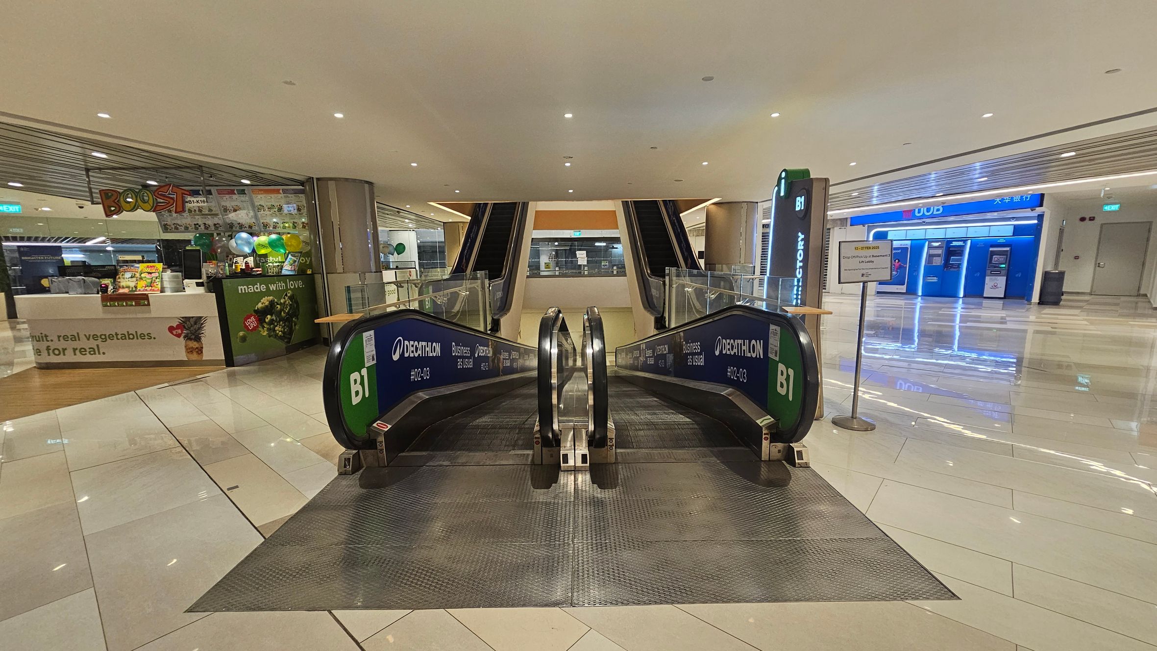 In the foreground, there are two escalators leading downwards, with green signage indicating "B1" and "Decathlon". On the left, there's a shop with a "Boost" sign, featuring a counter and colourful balloons. On the right, there is a bank area with several ATMs under a blue "UOB" sign. The floor is shiny and tiled, reflecting the ceiling lights. The ceiling is smooth with recessed lighting. In the foreground, there are two escalators leading downwards, with green signage indicating "B1" and "Decathlon". On the left, there's a shop with a "Boost" sign, featuring a counter and colourful balloons. On the right, there is a bank area with several ATMs under a blue "UOB" sign. The floor is shiny and tiled, reflecting the ceiling lights. The ceiling is smooth with recessed lighting.