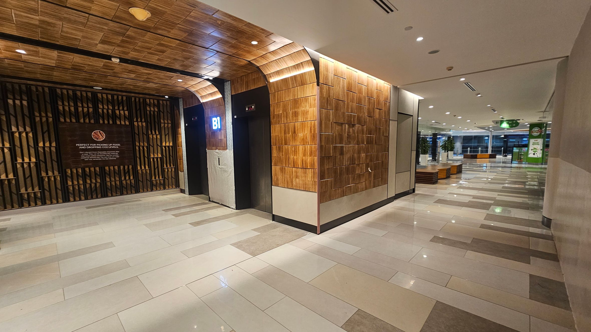 On the left, there is a wall with a wooden panel design and a sign with text. An elevator with a "B1" sign is next to it. The ceiling has a matching wooden pattern. On the right, the corridor extends with a tiled floor in shades of beige and grey. There are potted plants and seating areas along the corridor. The ceiling is white with recessed lighting. In the background, there are large windows and a green sign. On the left, there is a wall with a wooden panel design and a sign with text. An elevator with a "B1" sign is next to it. The ceiling has a matching wooden pattern. On the right, the corridor extends with a tiled floor in shades of beige and grey. There are potted plants and seating areas along the corridor. The ceiling is white with recessed lighting. In the background, there are large windows and a green sign.