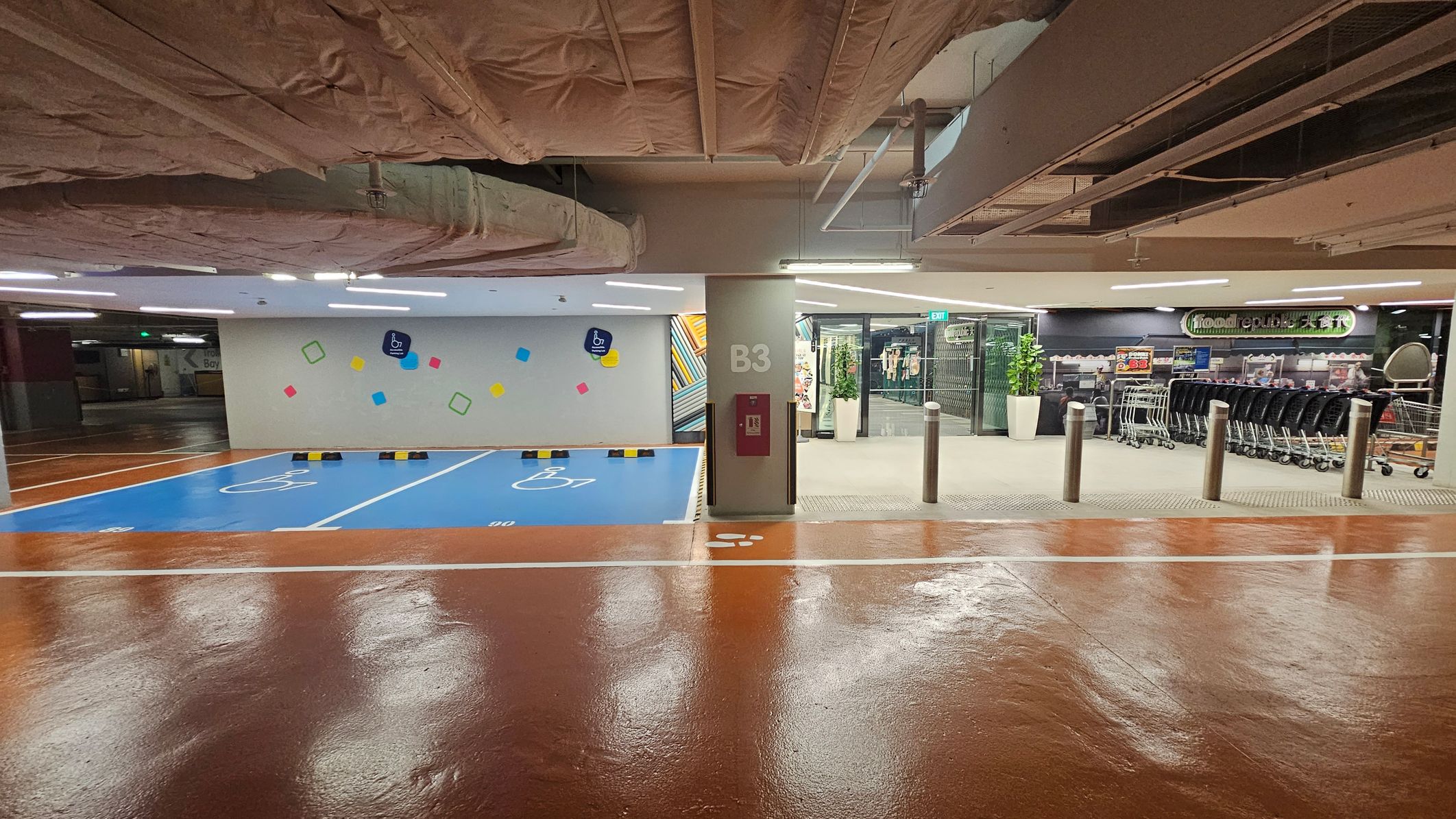 On the left, there are two blue parking spaces marked with wheelchair symbols, indicating accessibility. The wall behind is decorated with colourful geometric shapes. In the centre, a pillar marked "B3" has a fire hose cabinet attached. On the right, there is a supermarket entrance with shopping trolleys lined up outside. The floor is shiny and brown, and the ceiling has exposed ductwork and pipes. On the left, there are two blue parking spaces marked with wheelchair symbols, indicating accessibility. The wall behind is decorated with colourful geometric shapes. In the centre, a pillar marked "B3" has a fire hose cabinet attached. On the right, there is a supermarket entrance with shopping trolleys lined up outside. The floor is shiny and brown, and the ceiling has exposed ductwork and pipes.