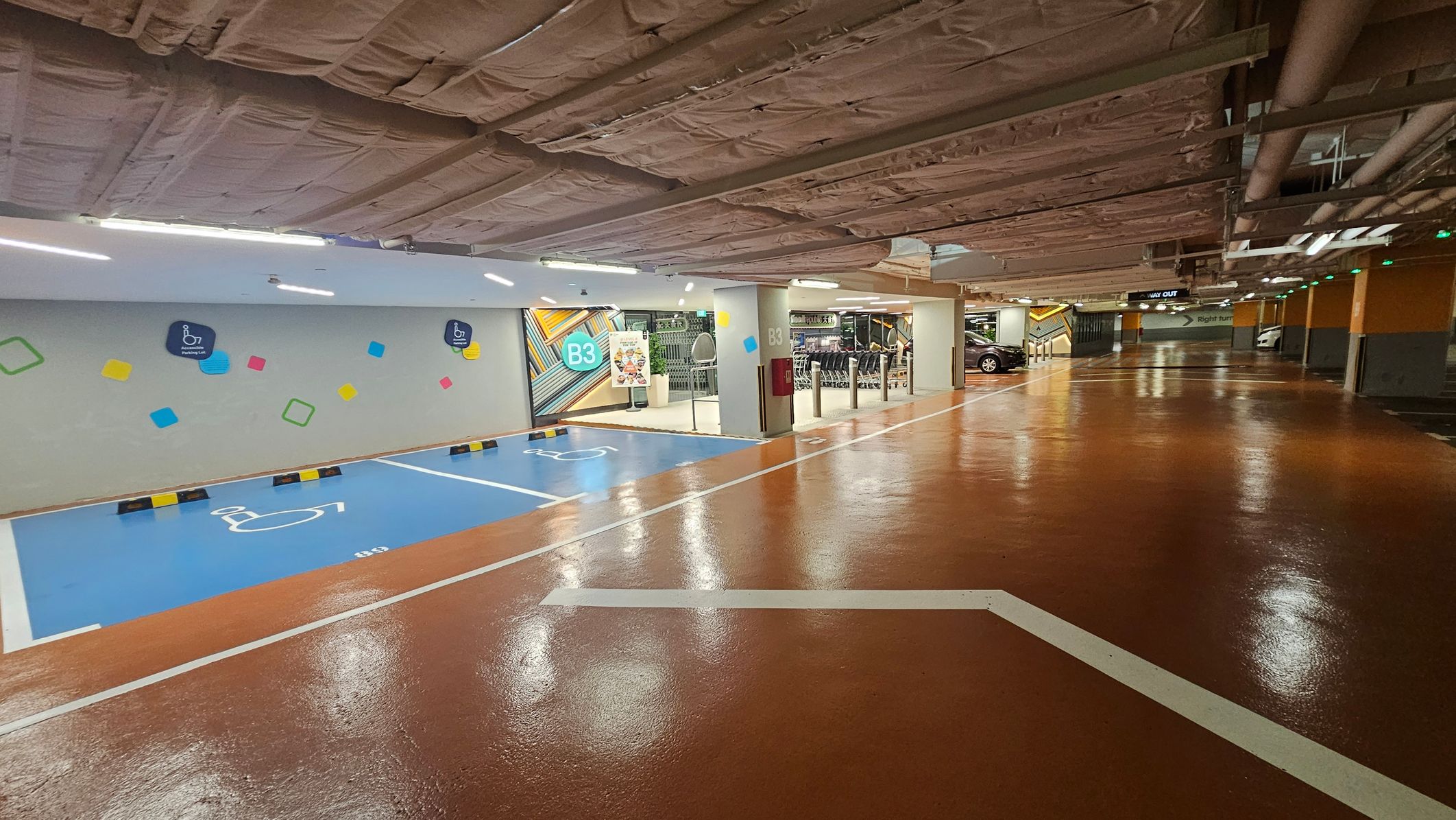 A parking garage with a shiny, brown floor and a ceiling covered in exposed insulation and pipes. On the left, there are blue parking spaces marked for disabled access, with colourful geometric shapes on the wall. In the middle, a sign indicates level B3, and there are shopping trolleys and a poster near the entrance. On the right, cars are parked, and a sign points to the way out. The area is well-lit with overhead lights. A parking garage with a shiny, brown floor and a ceiling covered in exposed insulation and pipes. On the left, there are blue parking spaces marked for disabled access, with colourful geometric shapes on the wall. In the middle, a sign indicates level B3, and there are shopping trolleys and a poster near the entrance. On the right, cars are parked, and a sign points to the way out. The area is well-lit with overhead lights.
